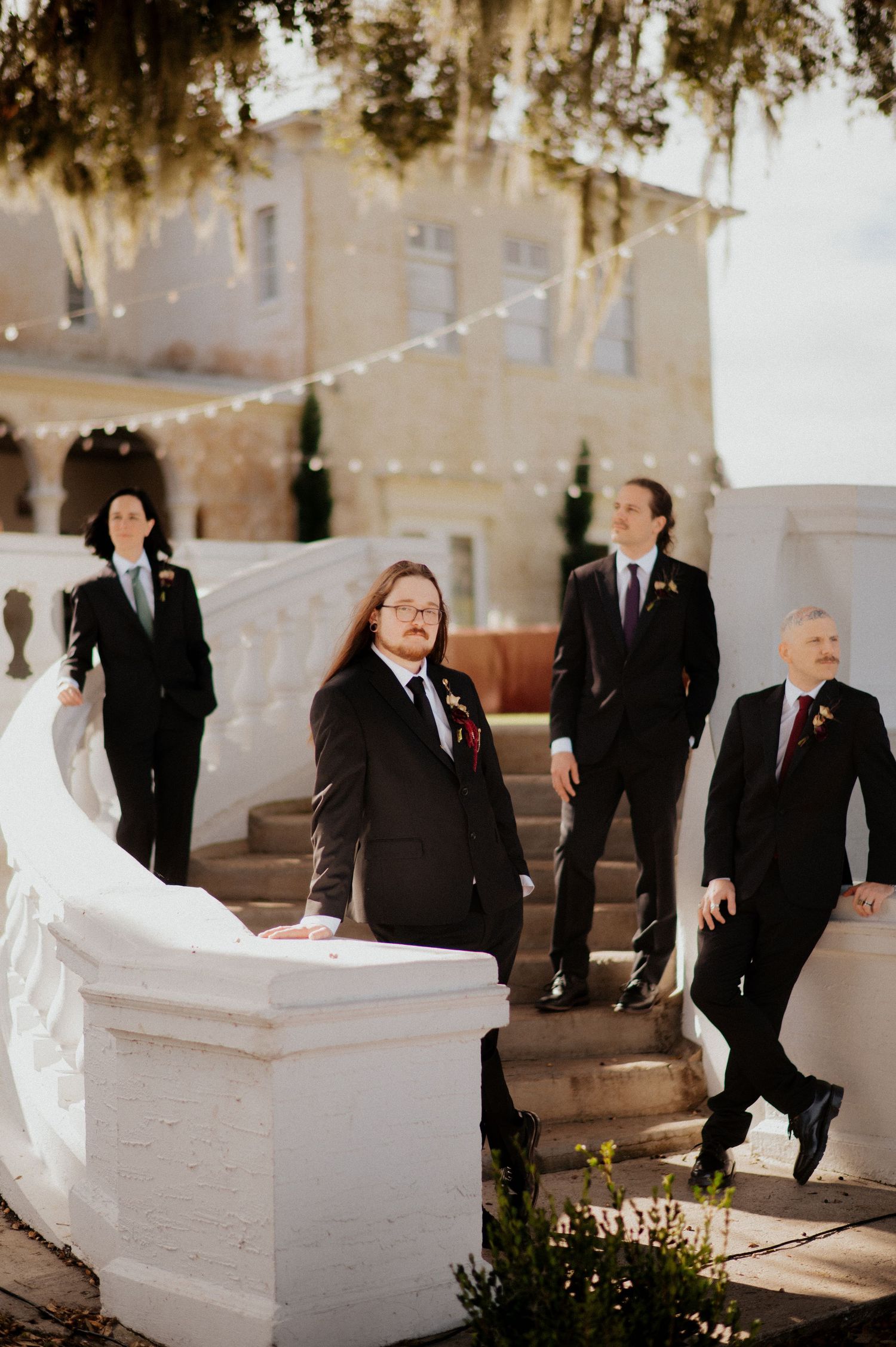 Wedding party members gathered around white stone benches in a courtyard with string lights overhead.