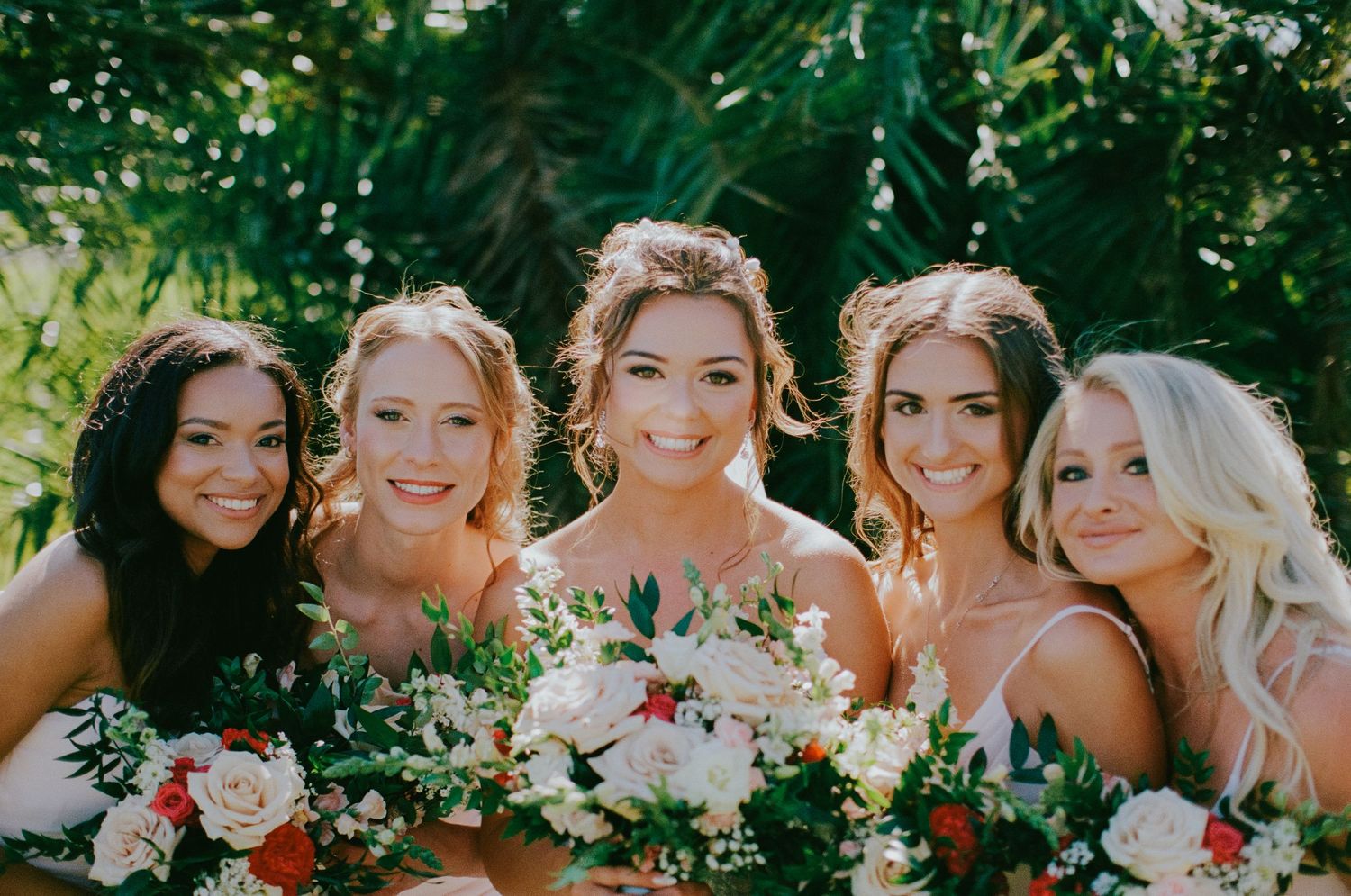 Five bridesmaids in white dresses smiling and holding lush floral bouquets surrounded by tropical greenery.