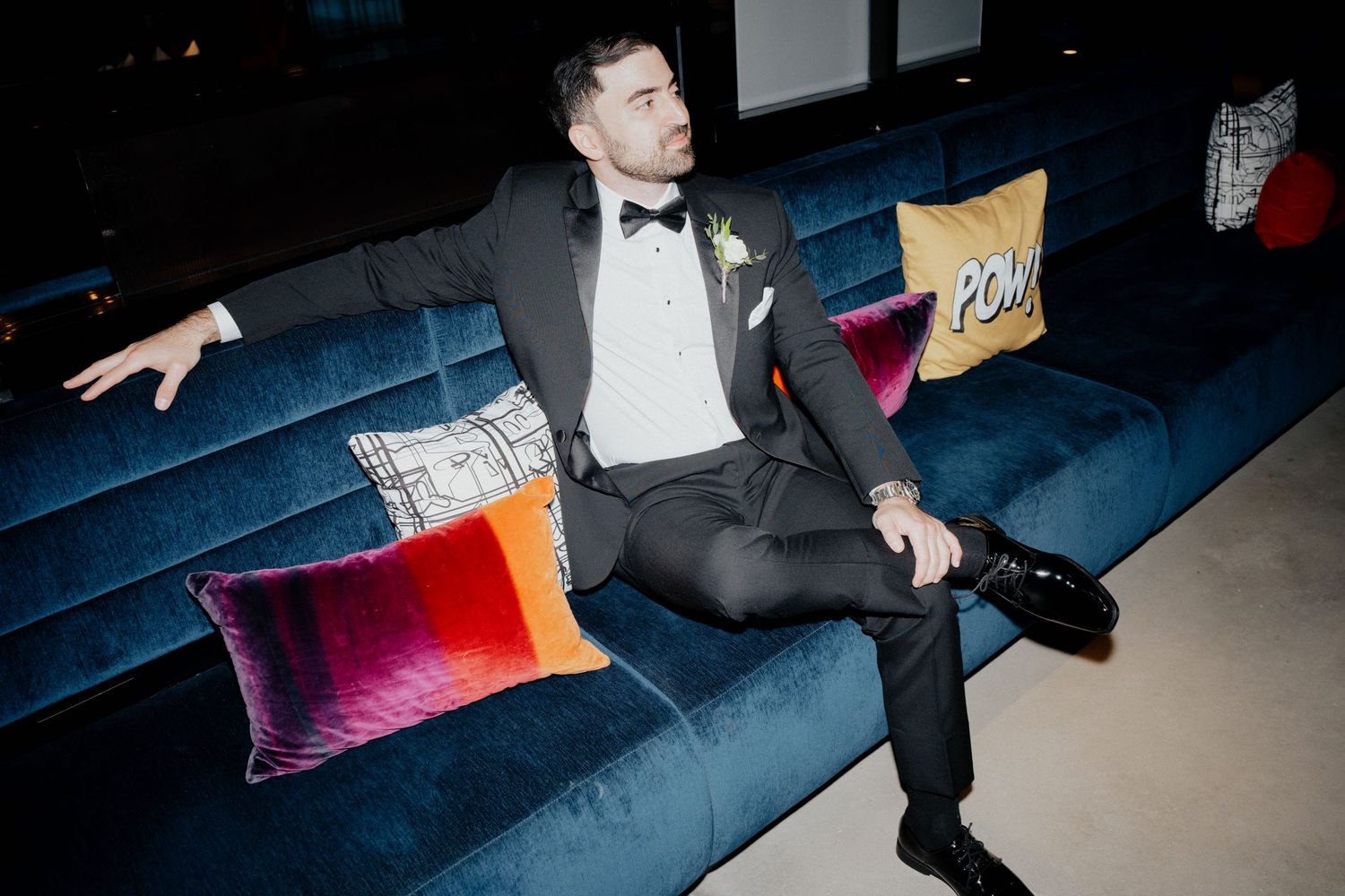 Stylish groom in black tuxedo relaxes on blue velvet sofa with colorful accent pillows.