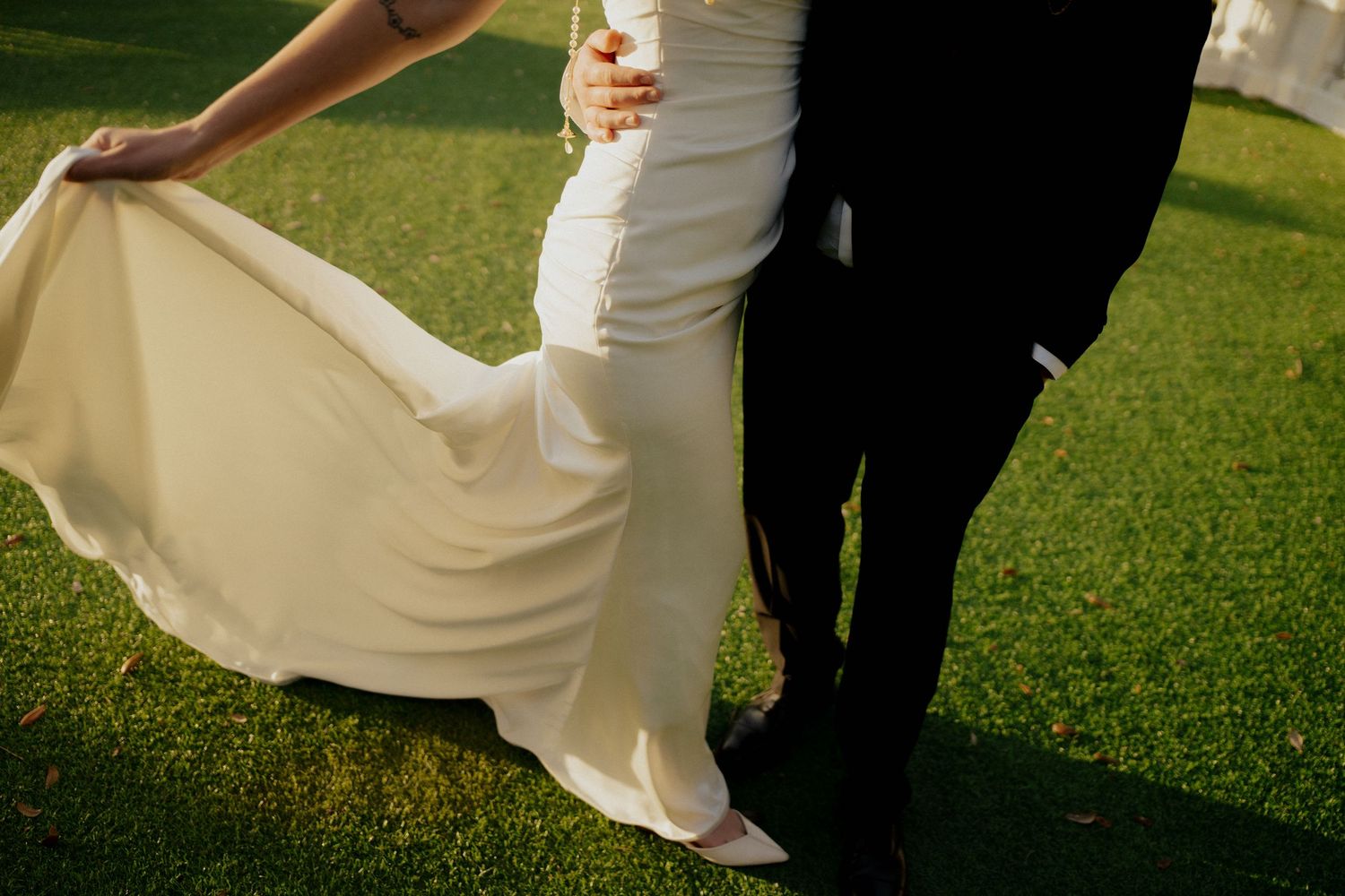 Bride in white wedding gown and groom in black suit standing on green grass during outdoor wedding ceremony.