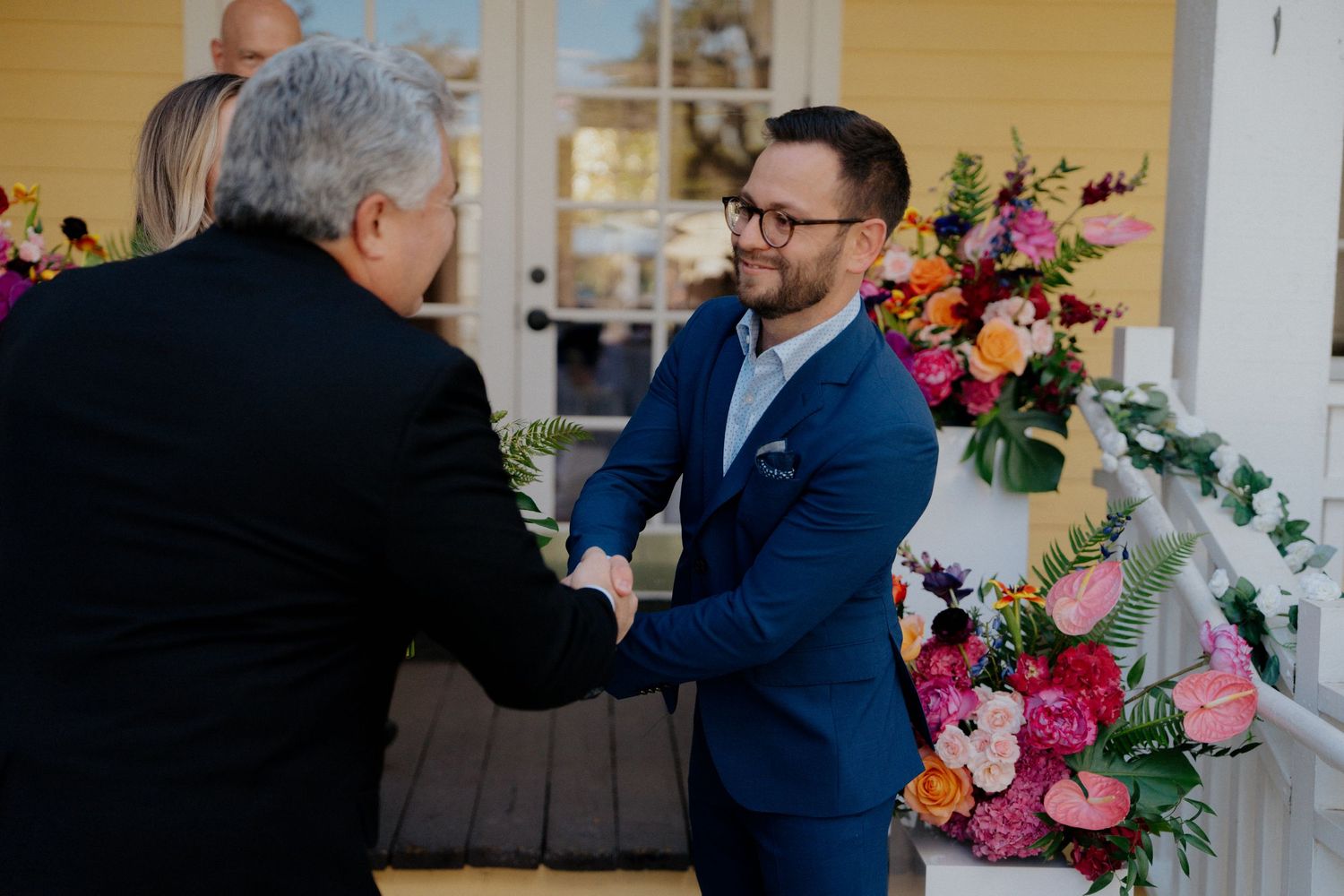 Two grooms in suits shake hands near colorful floral arrangements at an outdoor wedding ceremony.