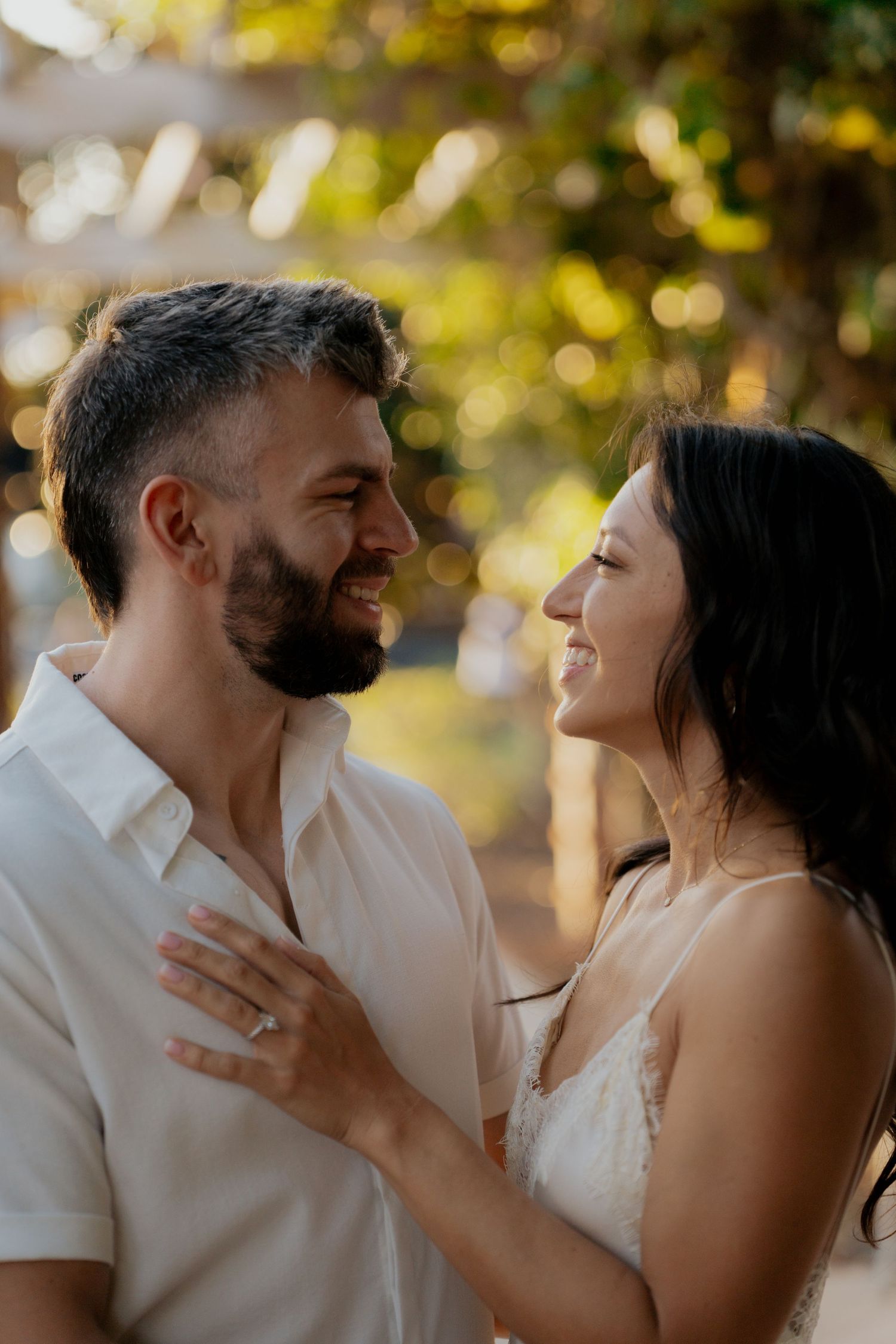Smiling engaged couple gazing at each other with bokeh garden background, woman wearing engagement ring.