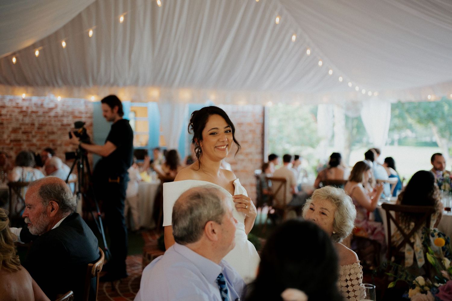 Wedding guests mingle and dance under a white tent during an evening reception celebration.