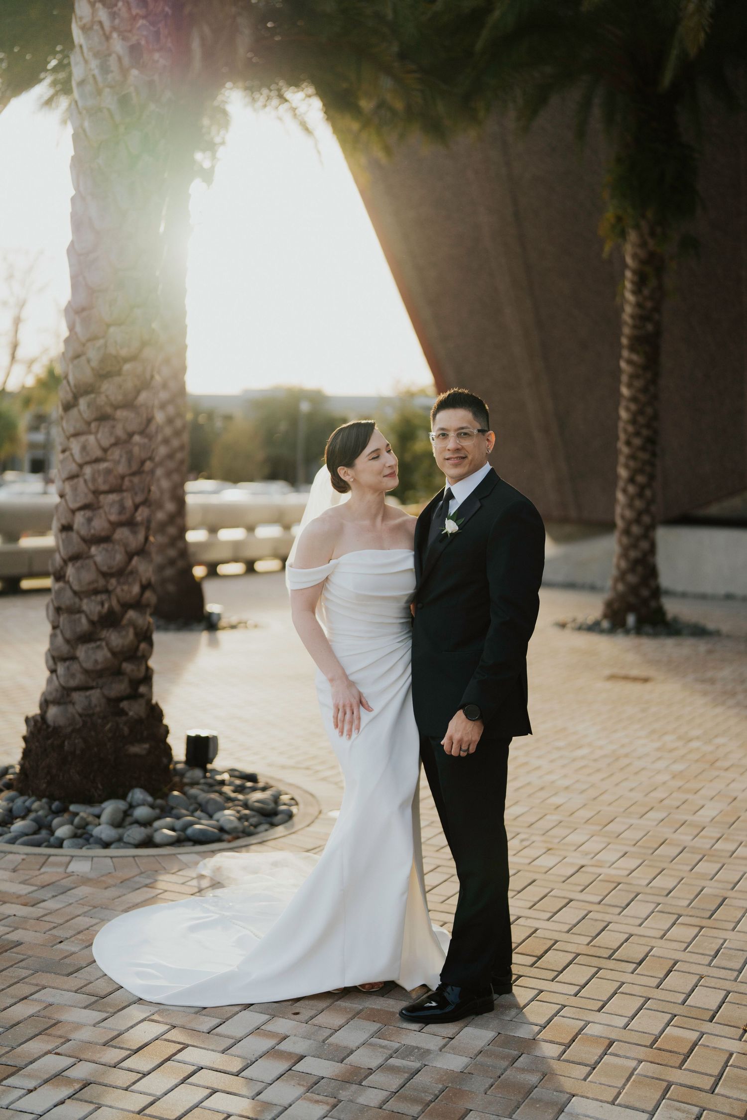 A couple in wedding attire poses together against a backdrop of palm trees during golden hour.