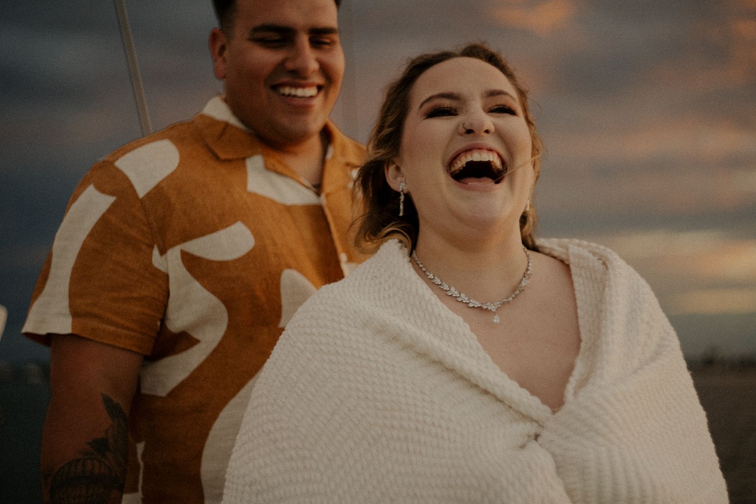 A joyful couple laughs together at sunset, woman wrapped in white blanket, man in brown patterned shirt.