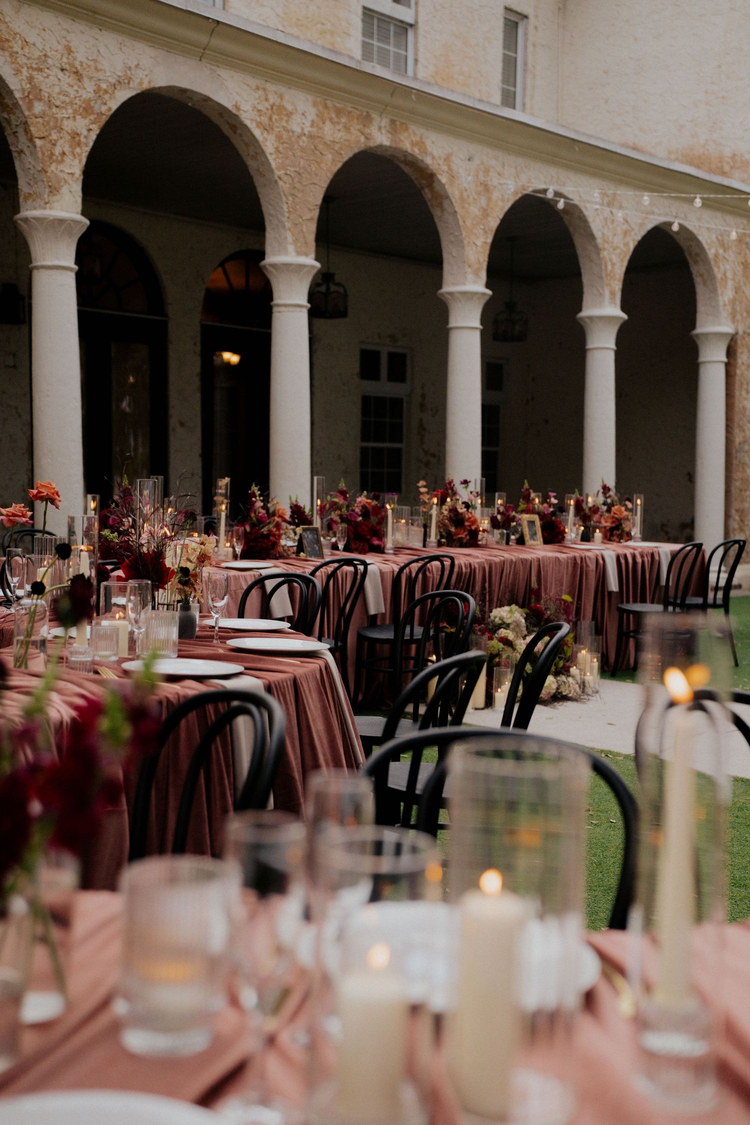 An elegant wedding reception setup with arched columns, burgundy tablecloths, and vintage chairs in a courtyard.