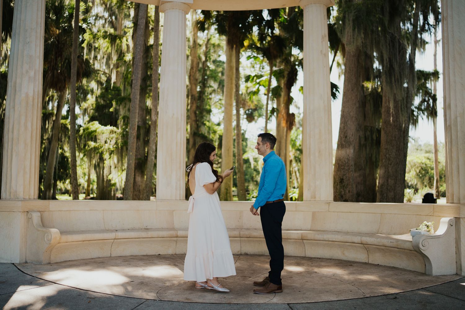 A couple stands beneath a white columned gazebo surrounded by tall trees in a park setting.