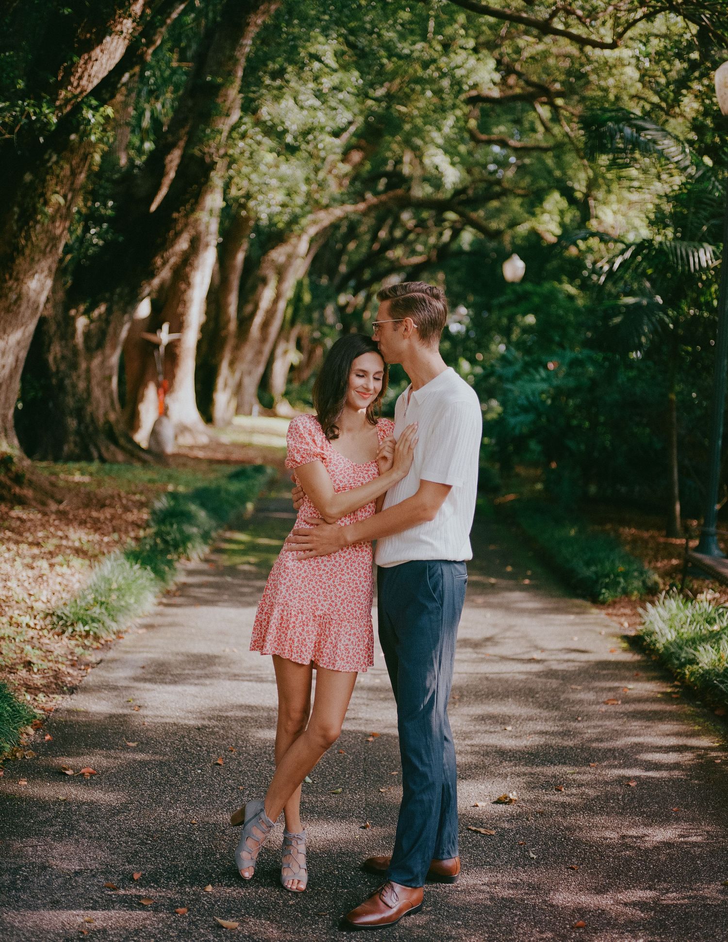 A couple shares a romantic moment on a tree-lined path during a summer photoshoot.