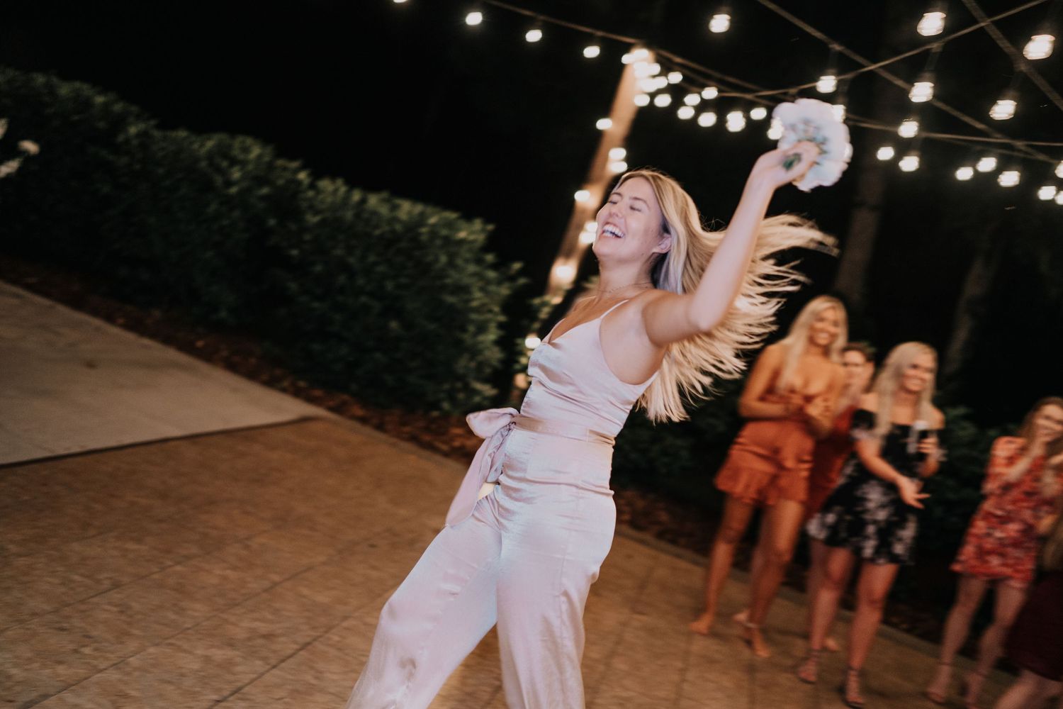 Bride in white jumpsuit tossing bouquet at outdoor wedding reception with string lights and guests.