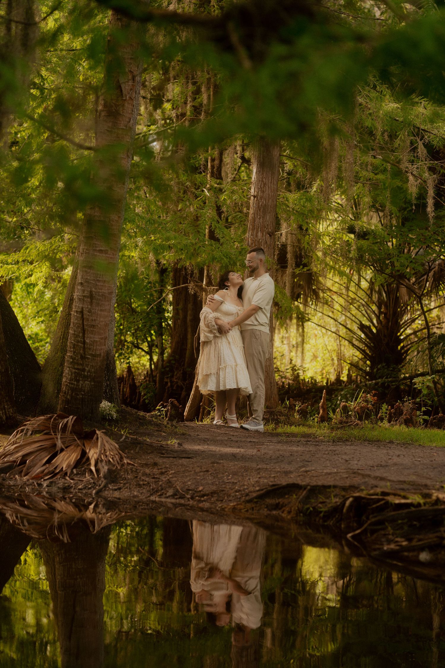 Couple embracing near tranquil pond in lush forest, their reflection mirrored in still water.