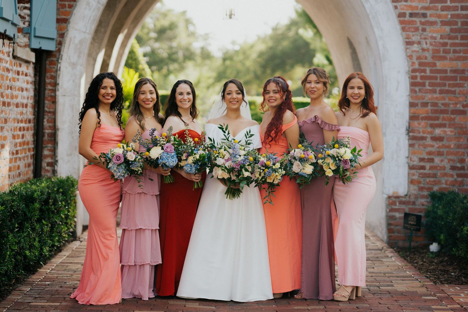 Wedding party in pink and coral dresses pose together under a brick archway.
