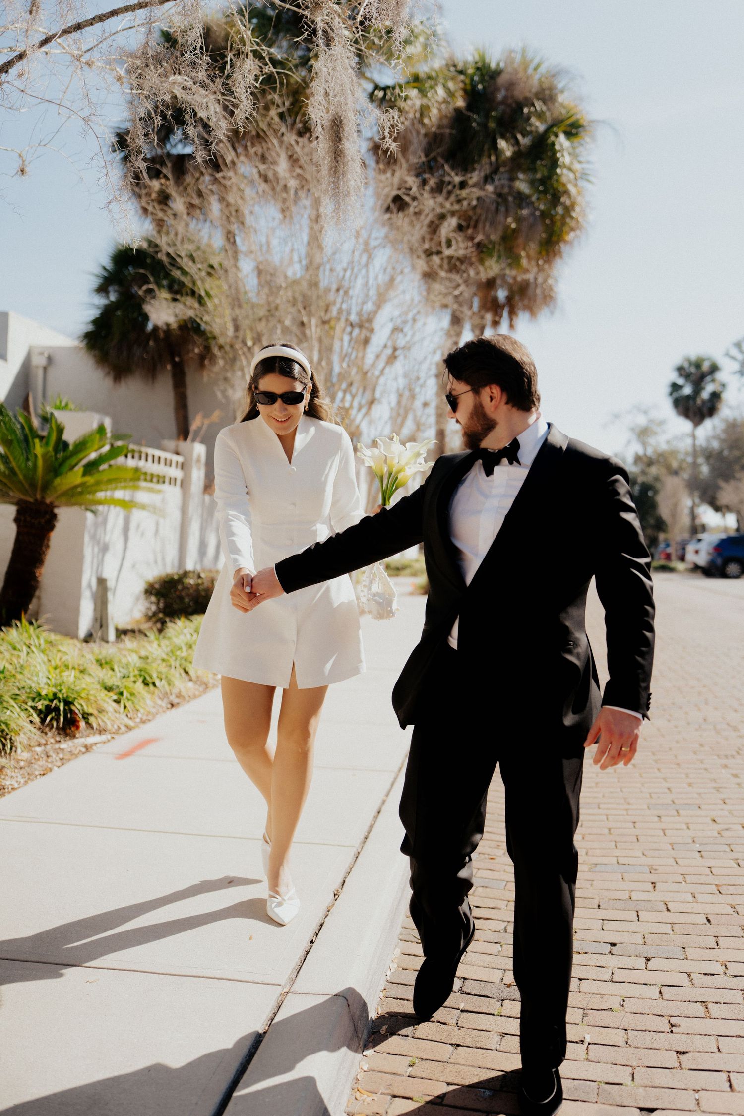 A couple in contrasting black and white outfits walking together on a palm tree-lined brick pathway in bright sunlight.