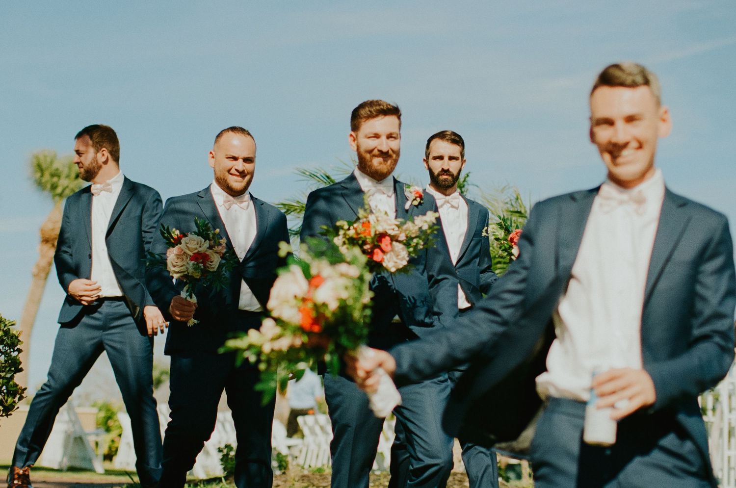 Groomsmen in navy suits holding colorful bouquets outdoors at a wedding ceremony on a sunny day.