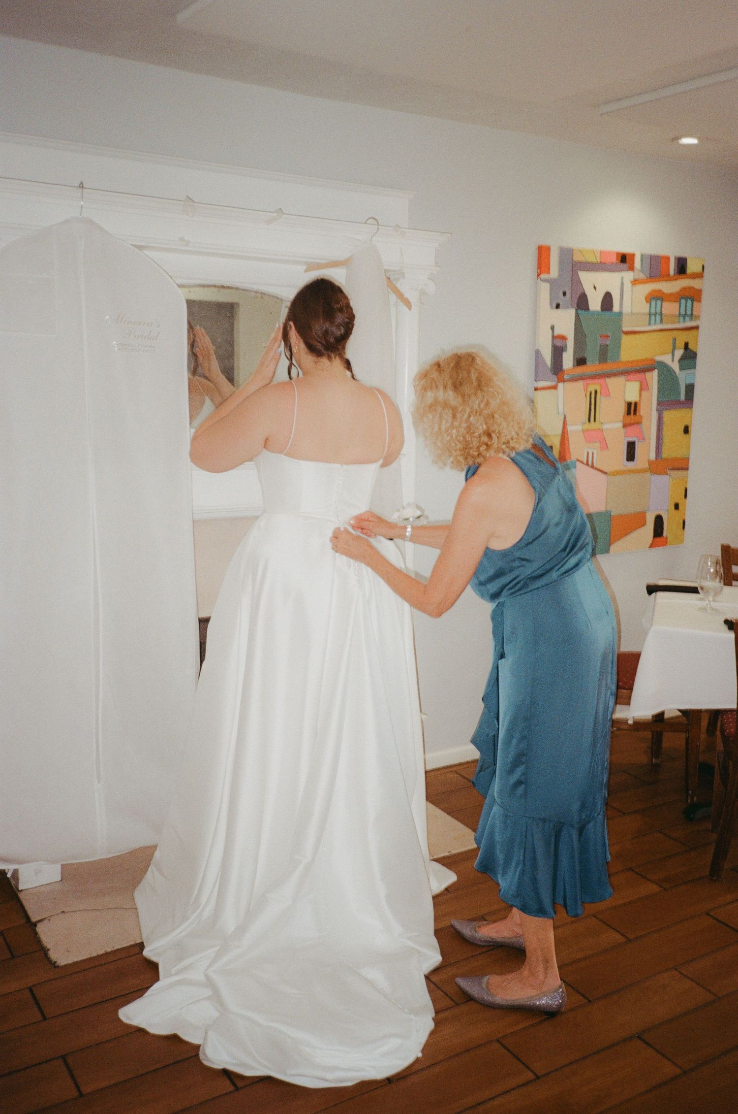 A bride in a white gown gets her dress fastened by a woman in a teal dress, standing before a mirror.