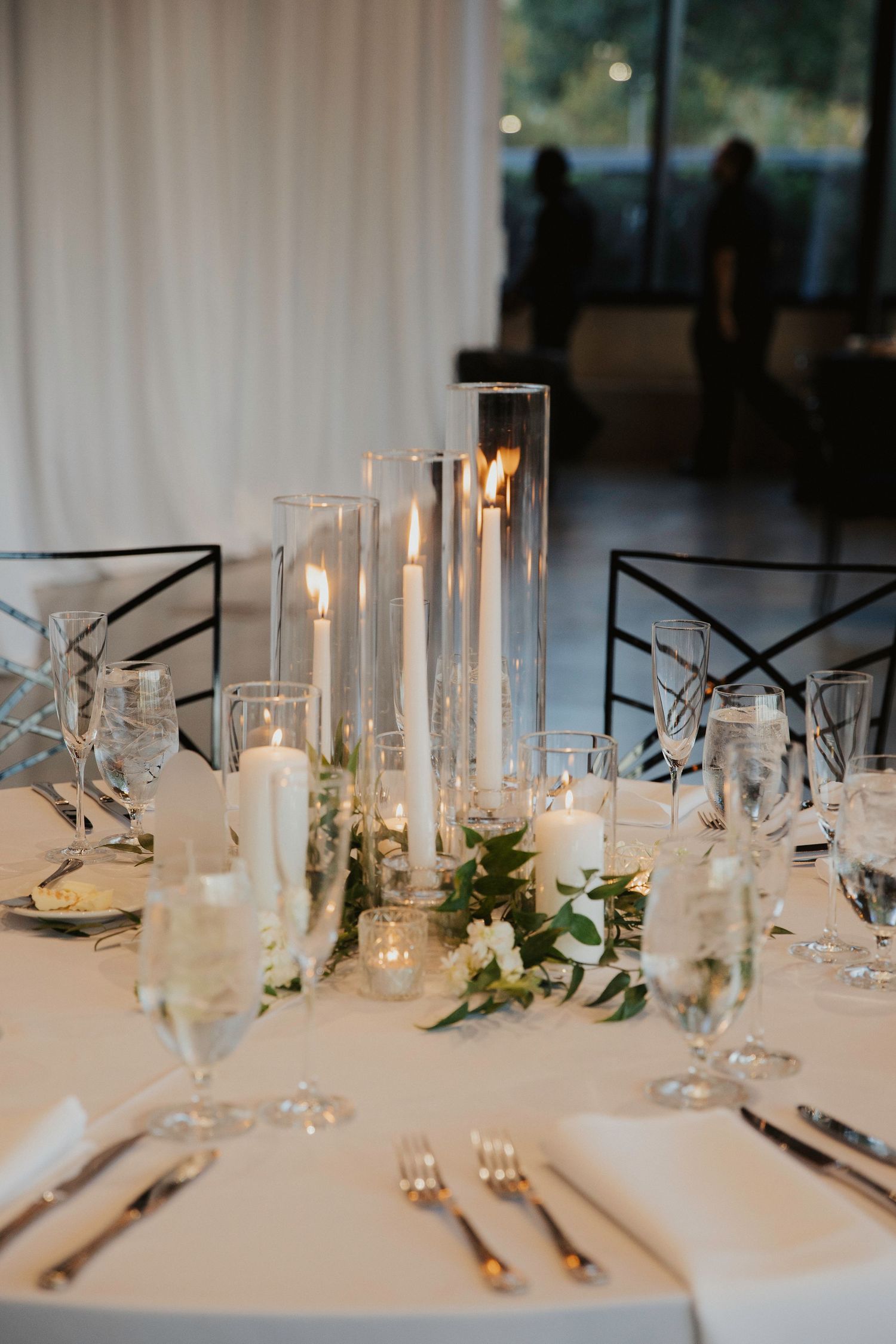 Elegant wedding reception table setting with tall glass candleholders and white floral accents on crisp linens.