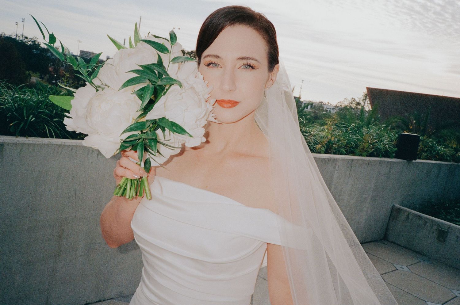 A bride in an elegant off-shoulder wedding dress holds a lush green and white bouquet against a natural outdoor backdrop.