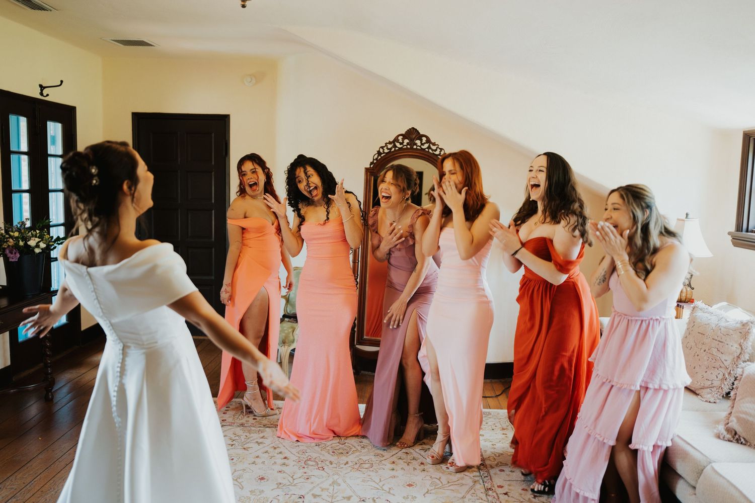 Bridesmaids in coral and pink dresses help the bride get ready in a bright indoor setting.
