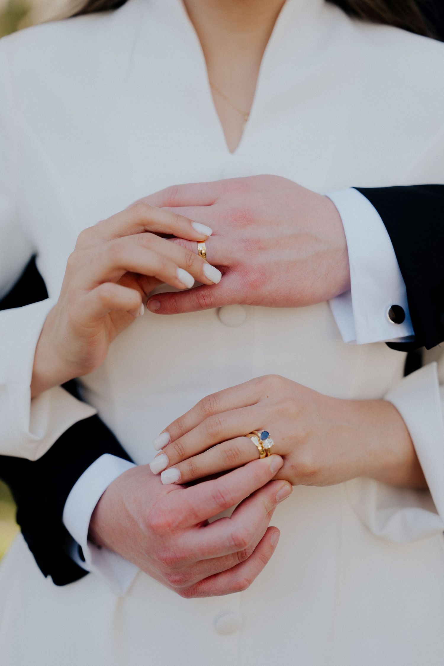 Close-up of newlyweds hands showing gold wedding rings and sapphire engagement ring on white dress.