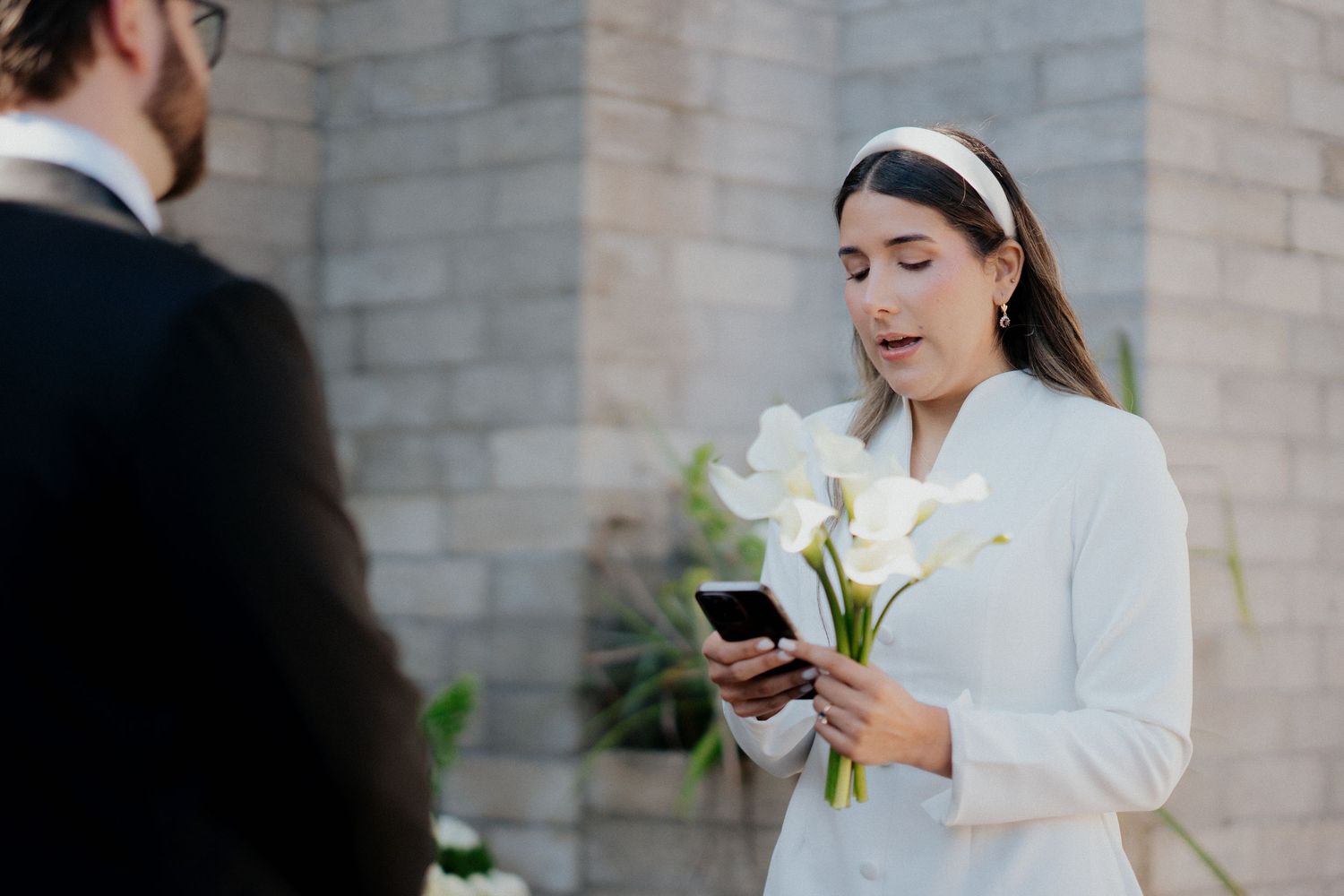Bride in white coat holds a glass of wine while standing outside a stone building in winter.
