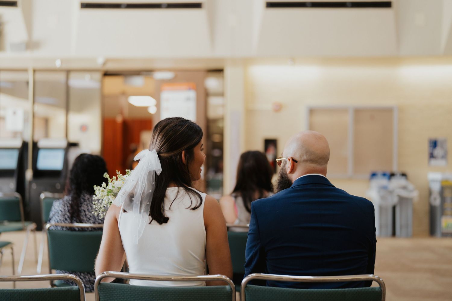 A couple sits side by side in white chairs at what appears to be a courthouse or office setting.