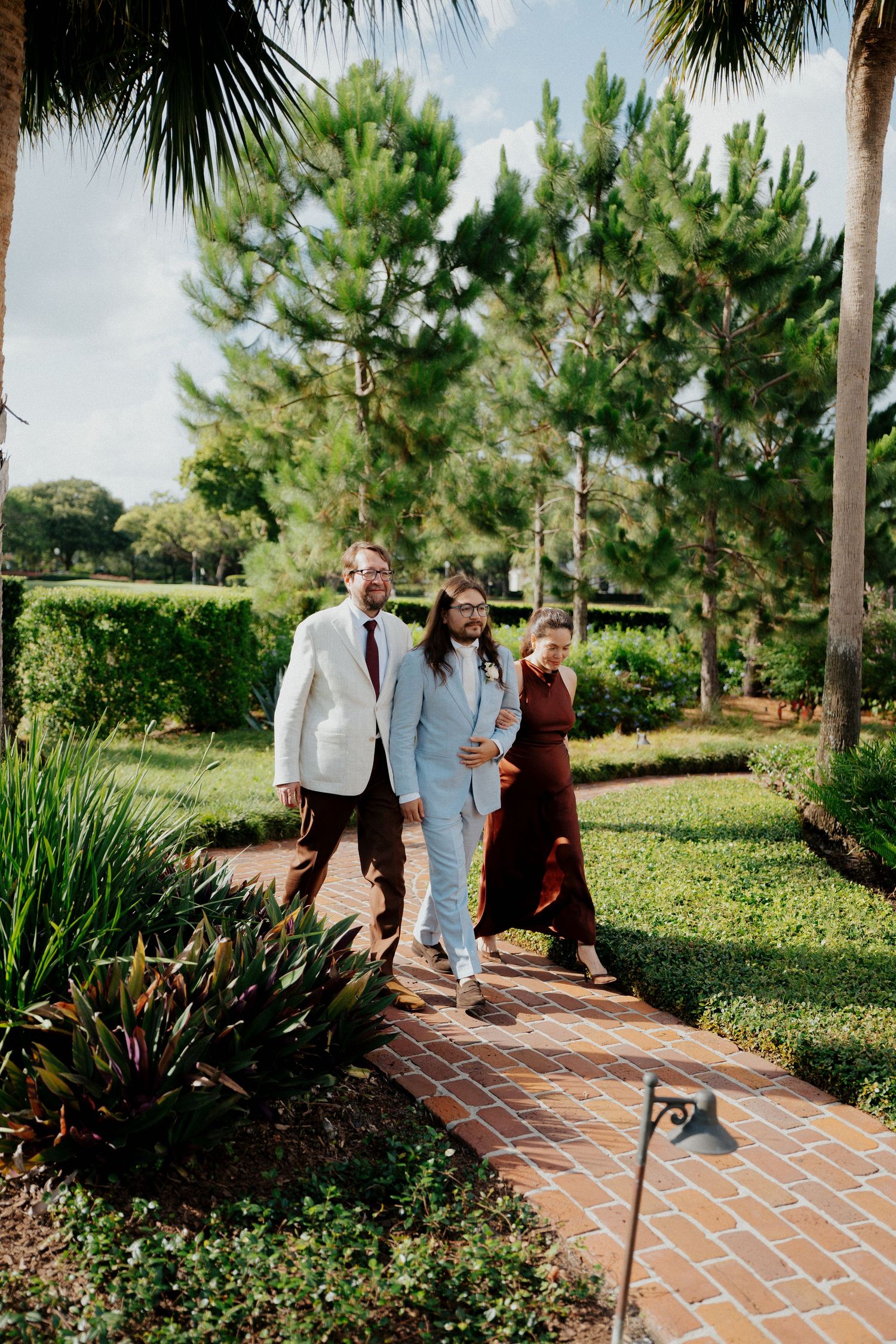 Wedding party walking along a scenic brick pathway lined with tropical plants and palm trees.
