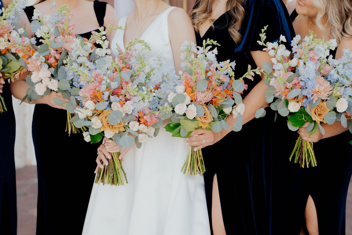 Wedding party stands in a row holding elegant pastel bouquets with blush and cream flowers against a neutral backdrop.