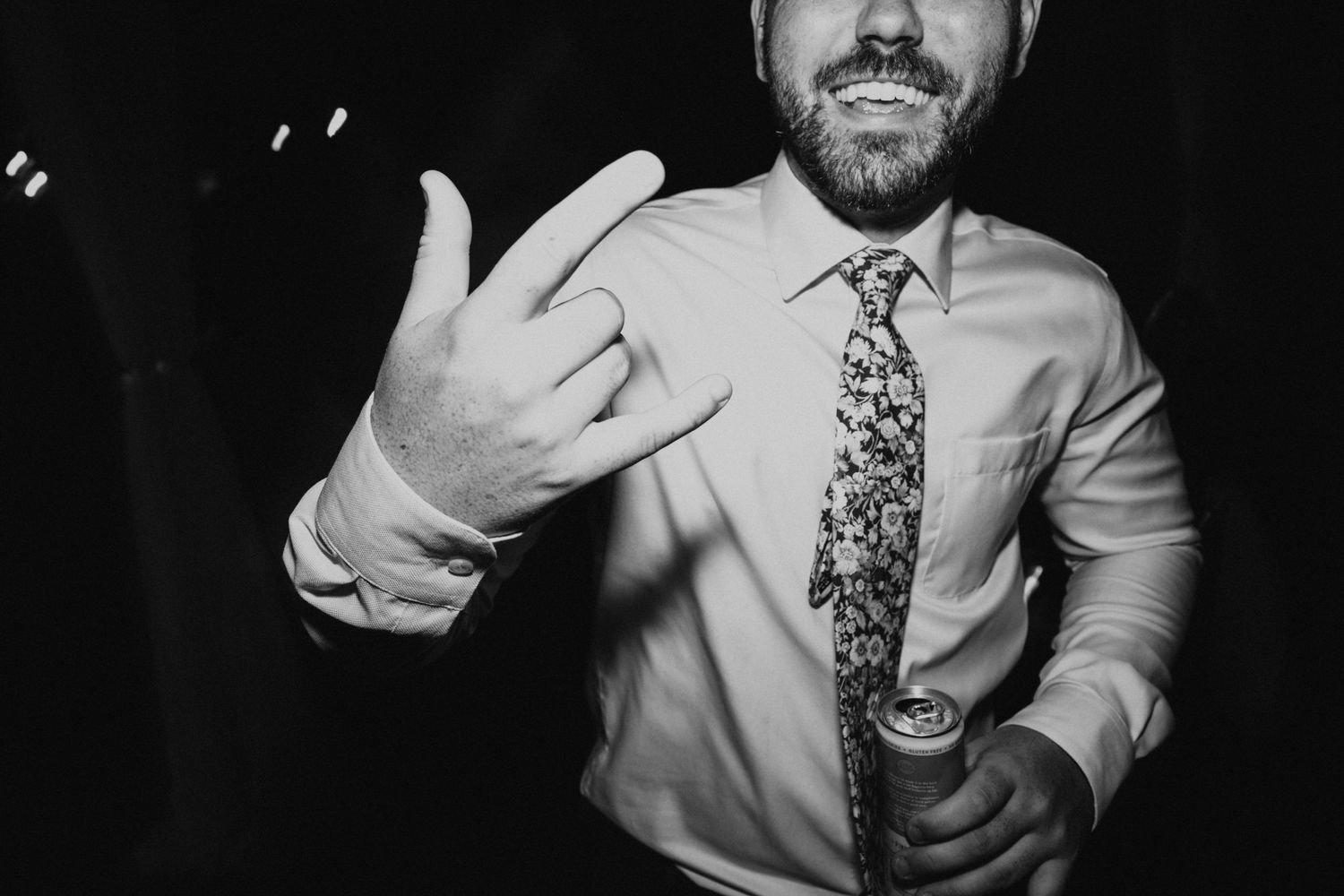 Groom in white shirt and floral tie making a rock on hand gesture while holding a drink at wedding reception.