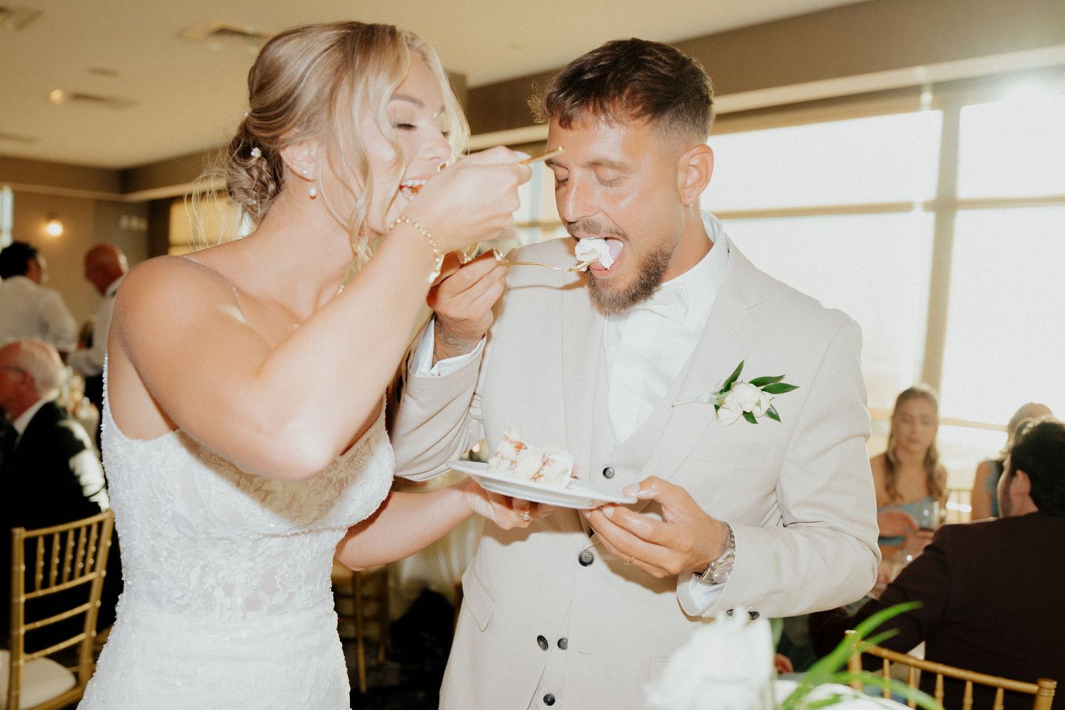 Bride and groom sharing a joyful cake-cutting moment at their wedding reception in a waterfront venue.