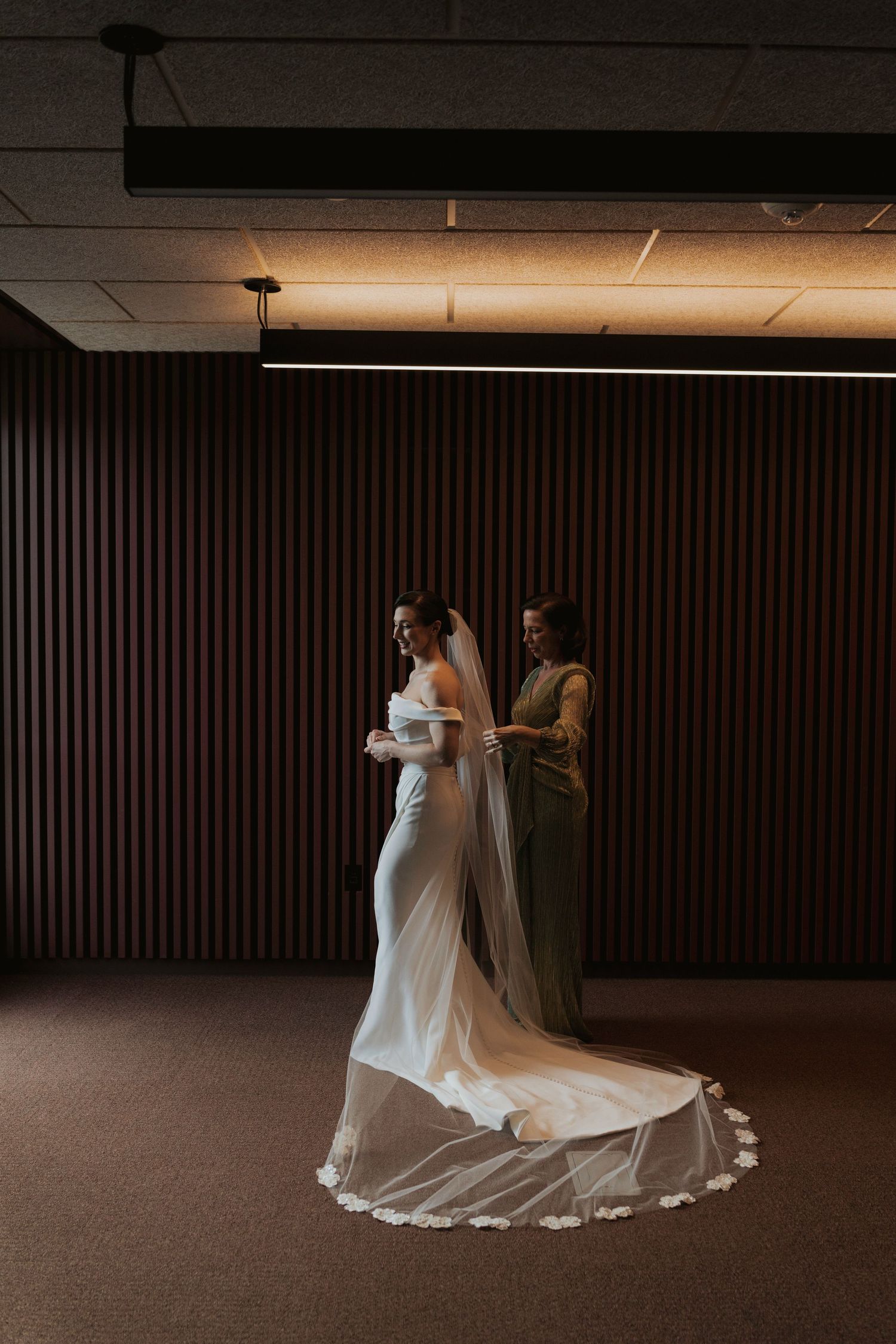 A couple in wedding attire poses in a dimly lit room with dramatic lighting and dark wood paneled walls.