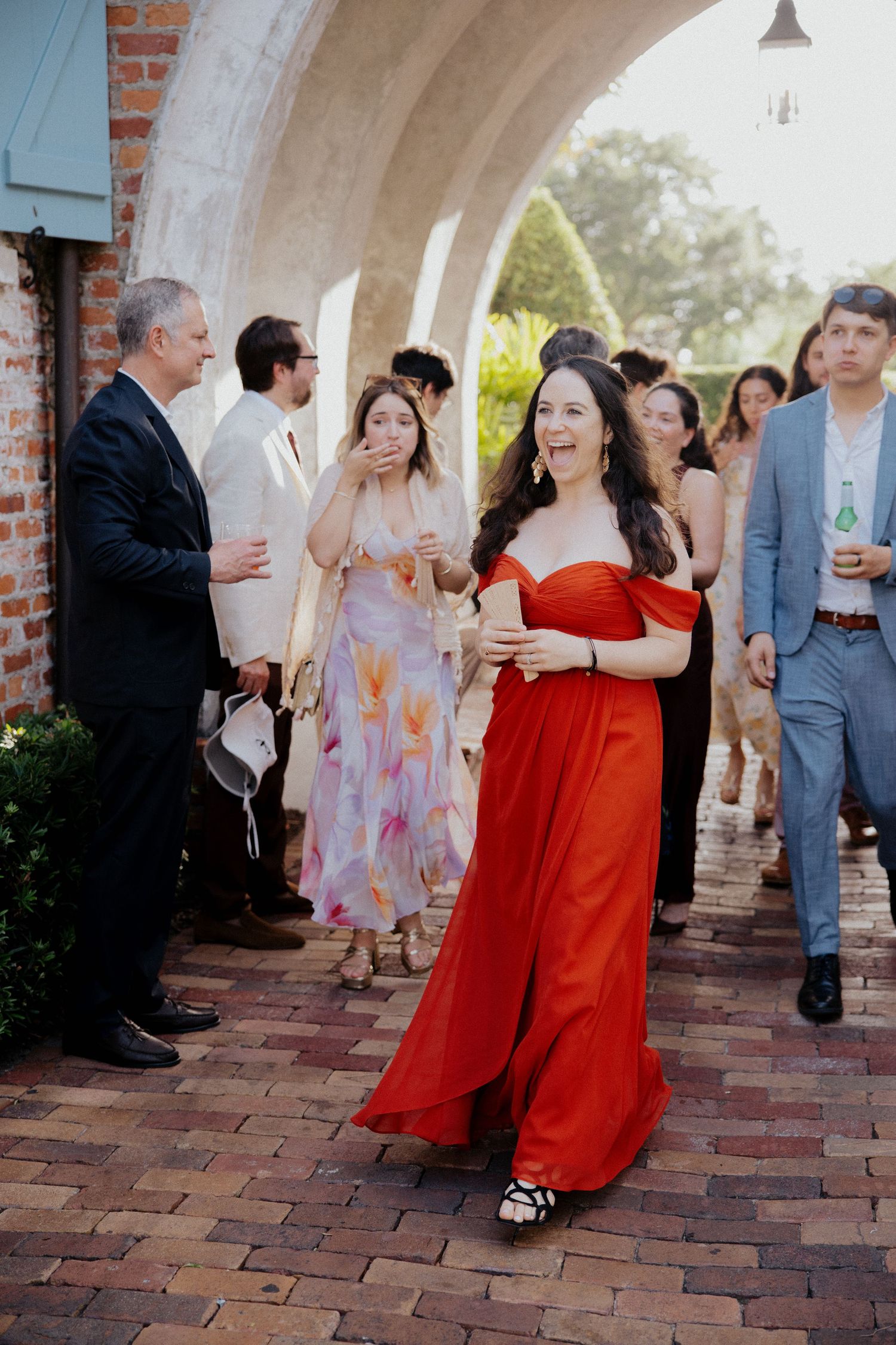 Wedding guests gather under a brick archway with a woman in a striking red jumpsuit laughing in the center.