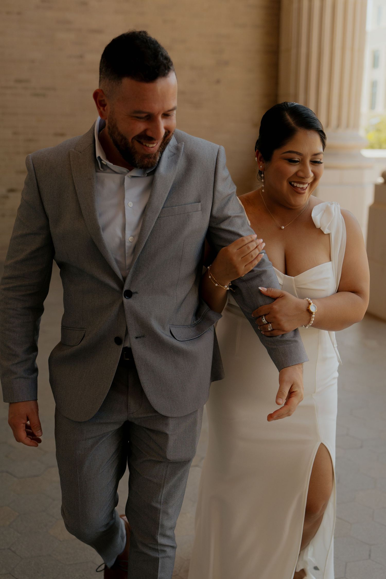 Smiling bride in white gown links arms with groom in gray suit between stone columns.