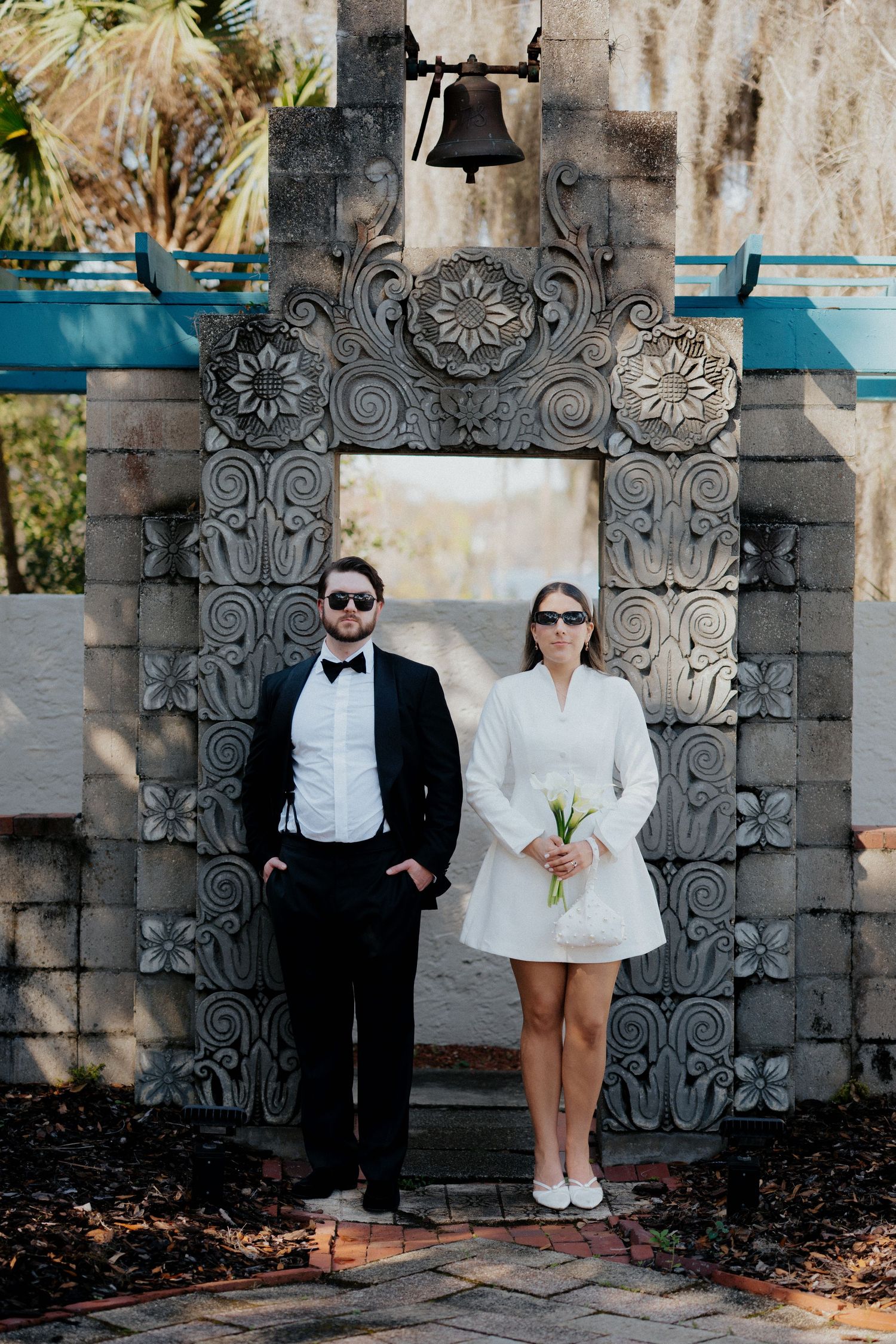 A couple in formal attire poses under an ornate stone archway with floral carvings.