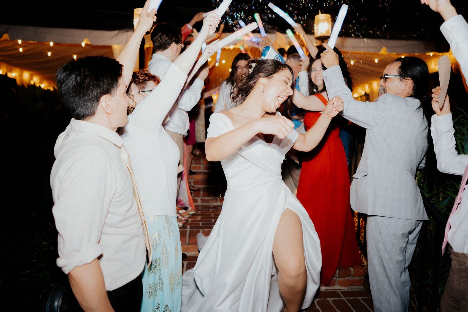 Energetic wedding reception dance party with guests raising their arms in celebration under ambient lighting.