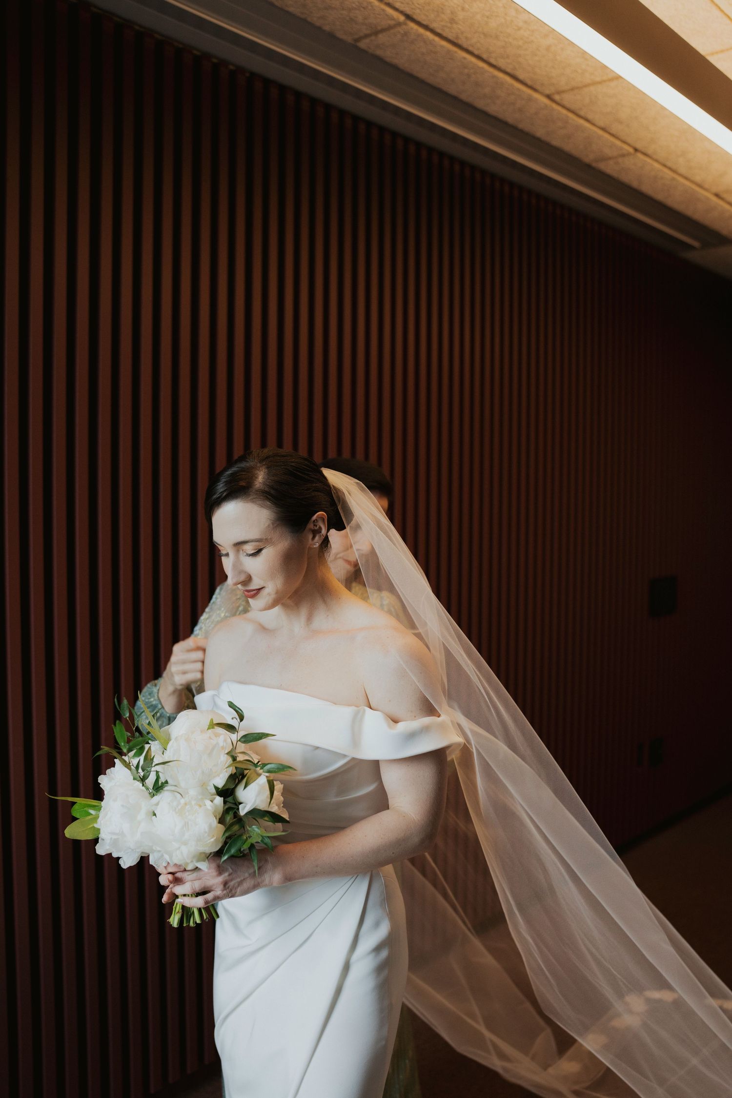 A bride in an elegant off-shoulder white gown holds a bouquet of white flowers against a wood-paneled wall.