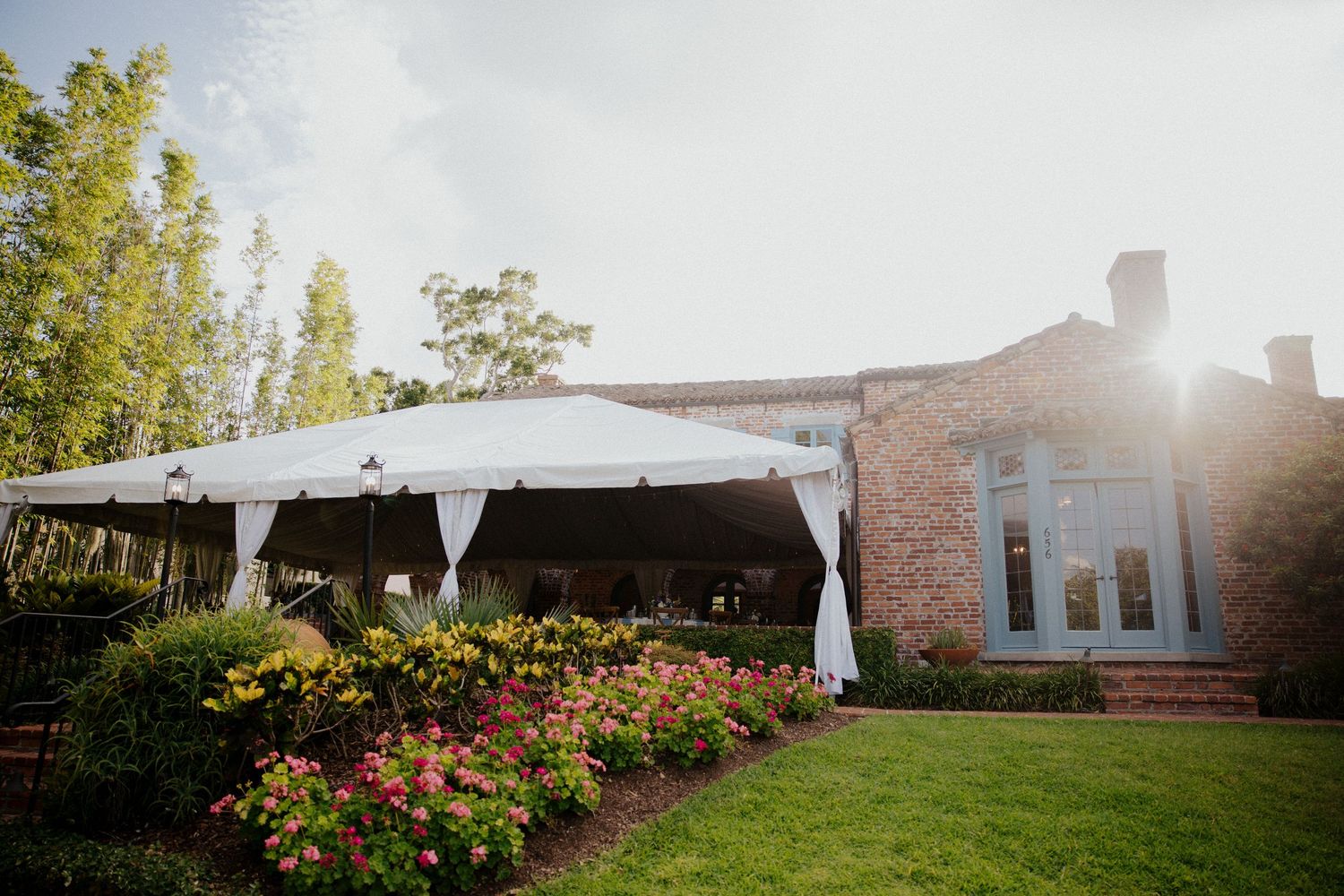 White wedding tent set up outdoors with sunlight streaming through trees and pink flowers in the garden.