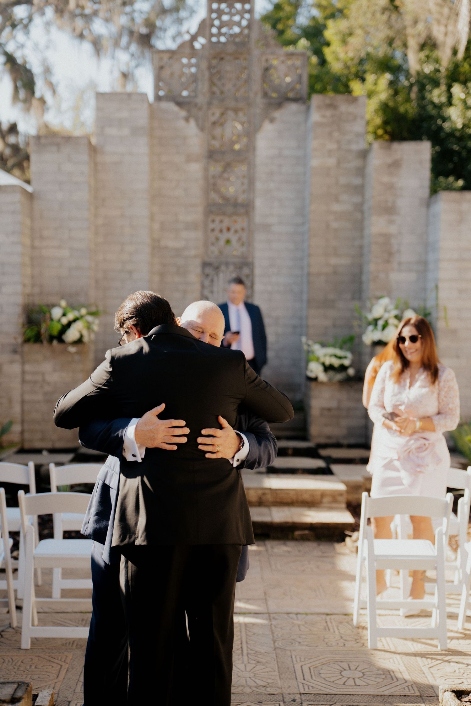 Emotional embrace between two people at a wedding ceremony with white chairs and decorative stone walls.