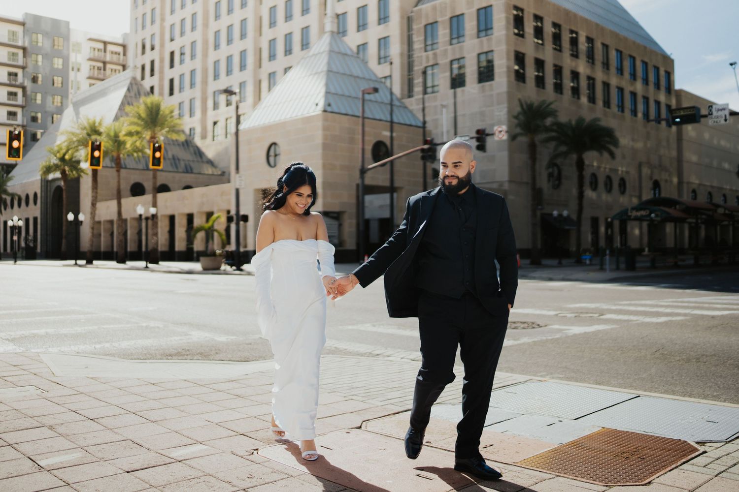 A couple in wedding attire walking hand in hand through a sunny urban street.
