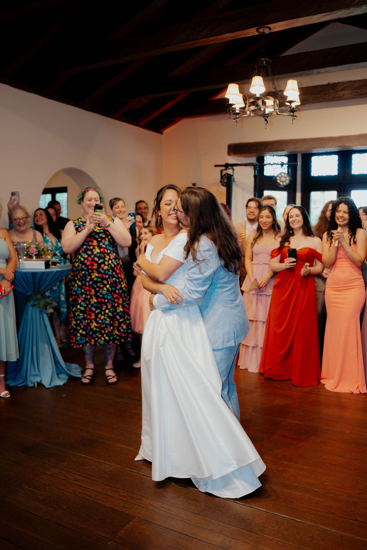Wedding reception guests dance together in colorful formal attire on a wooden dance floor under chandelier lighting.