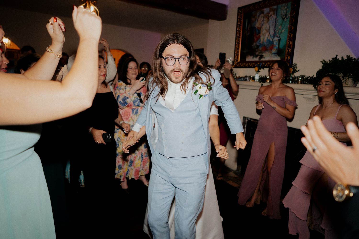 Wedding guests dance together during a lively reception celebration in a dimly lit venue with purple lighting.