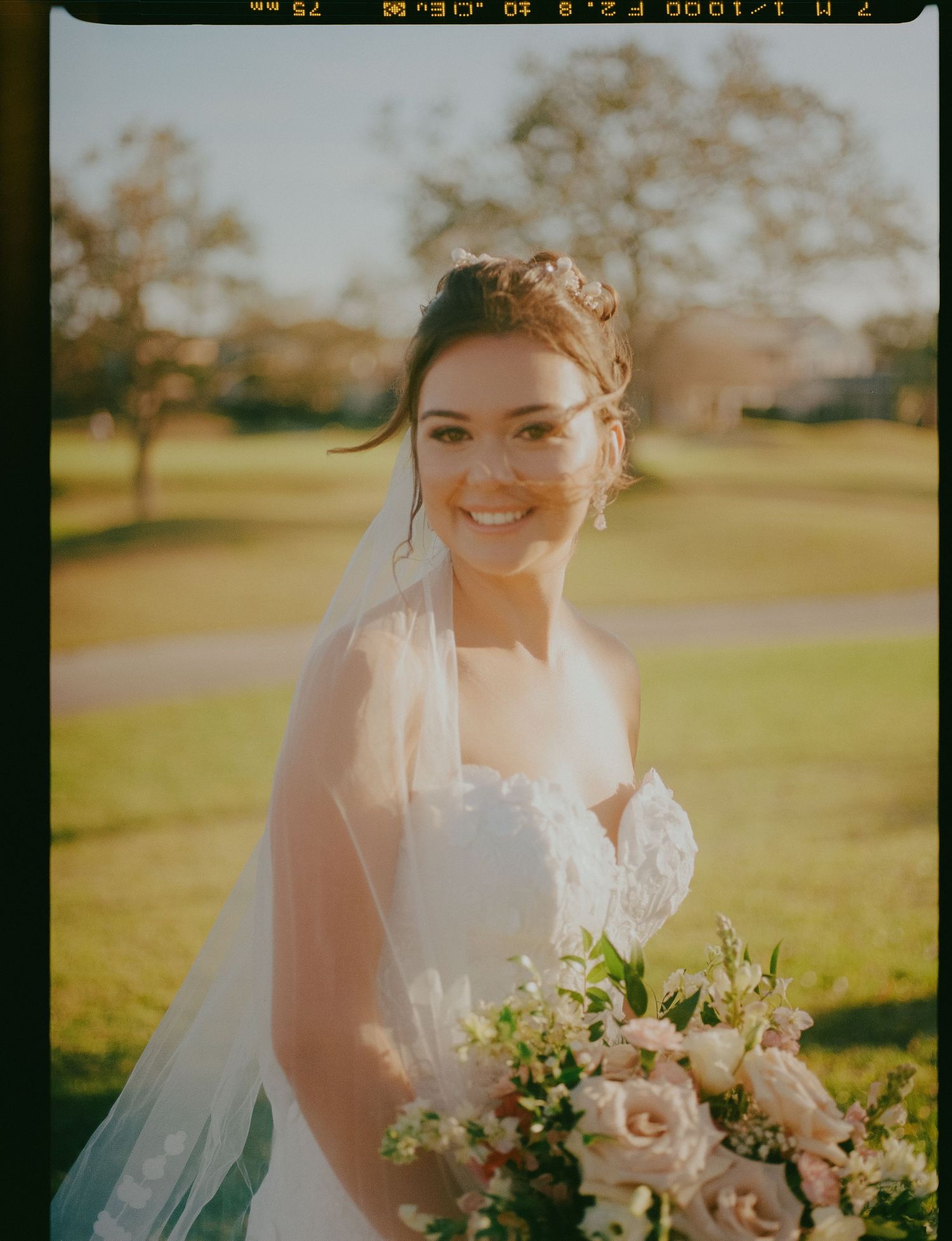 Smiling bride in a white strapless gown with veil holding a pink rose bouquet captured on film photography.
