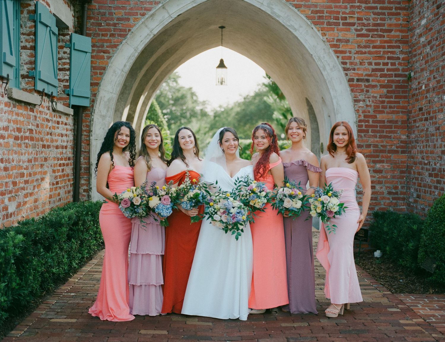 Bride with six bridesmaids in coral and mauve gowns holding bouquets under a brick archway.