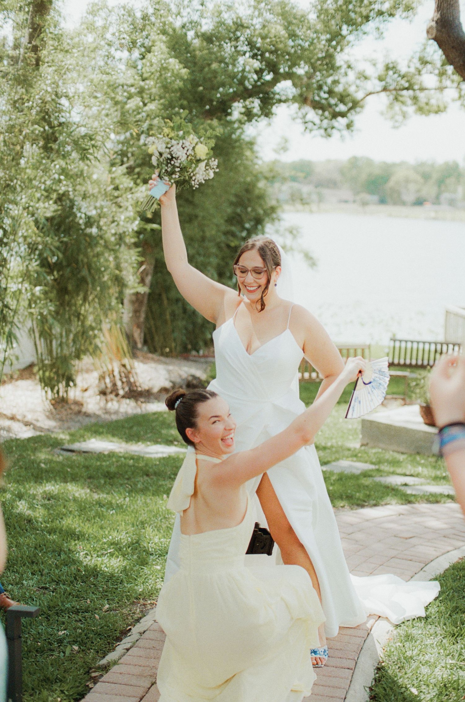 Two brides celebrate joyfully outdoors near a lake, one lifting the other while holding a bouquet on a sunny wedding day.
