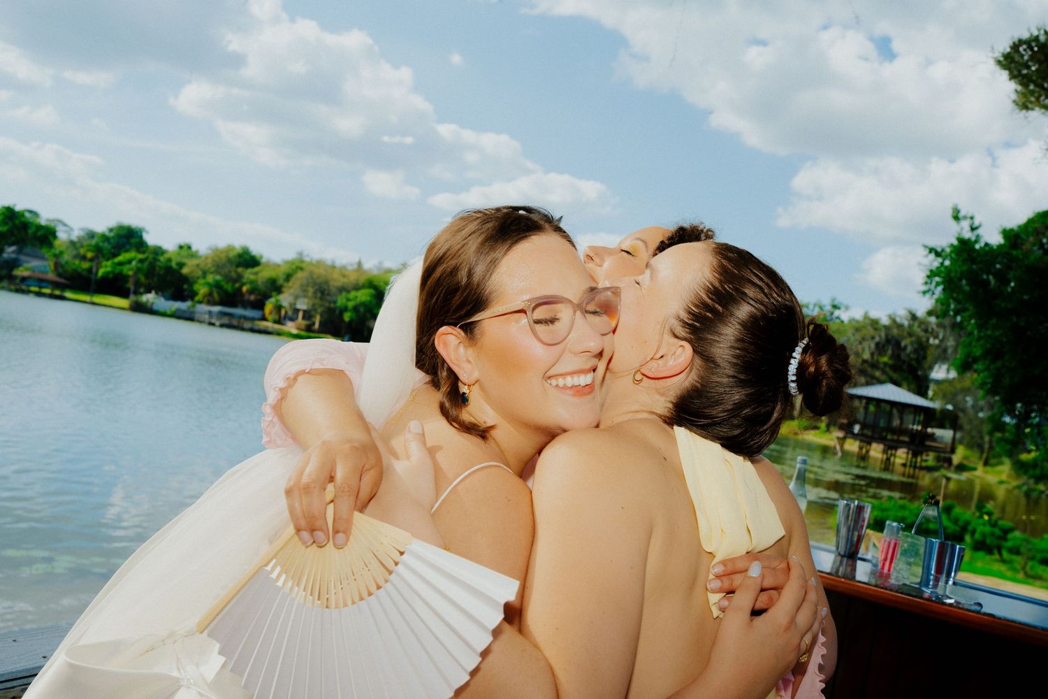 Two women hugging and smiling near a lake at an outdoor wedding celebration.