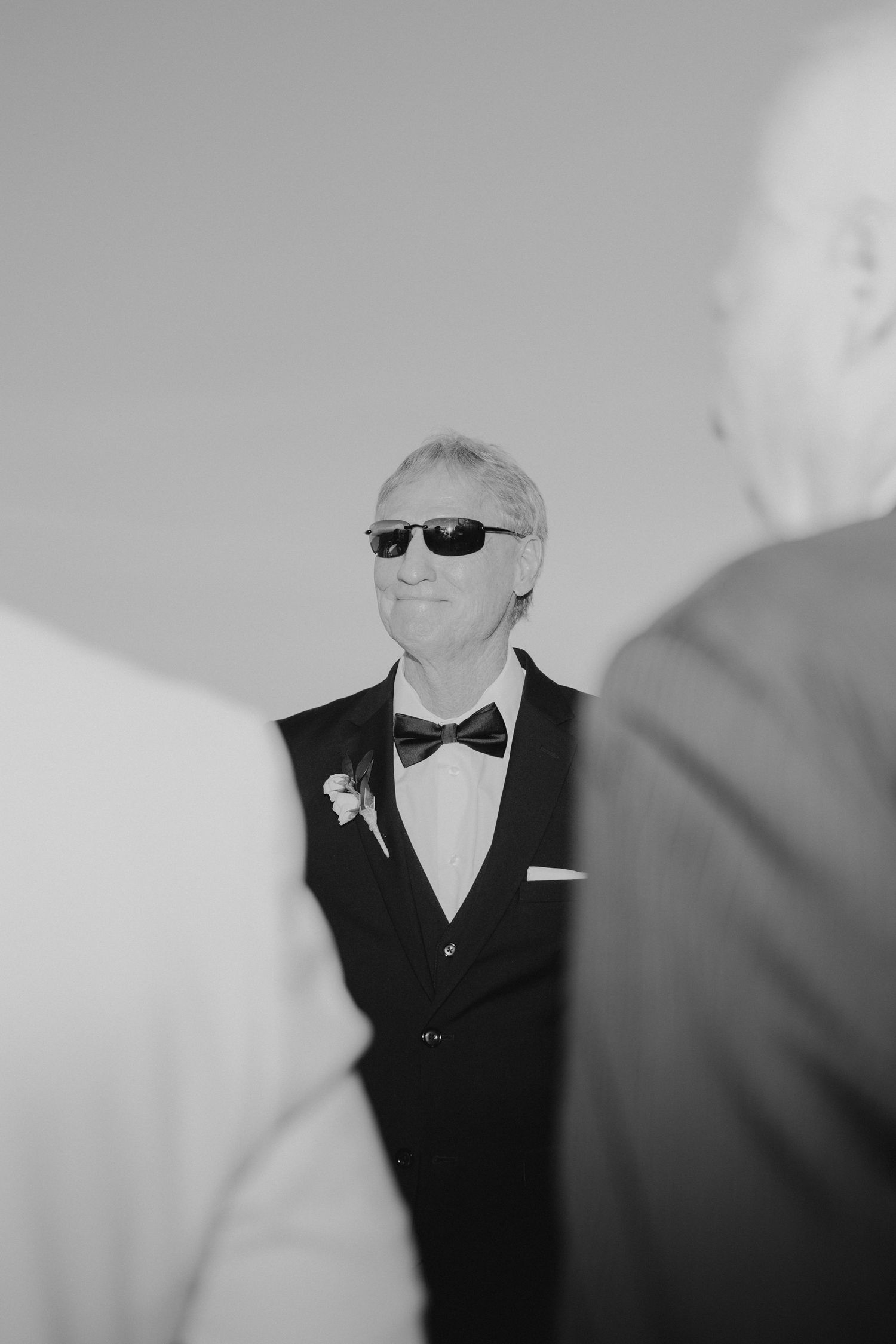 Black and white photo of a distinguished man in tuxedo and sunglasses wearing a boutonniere at a wedding.