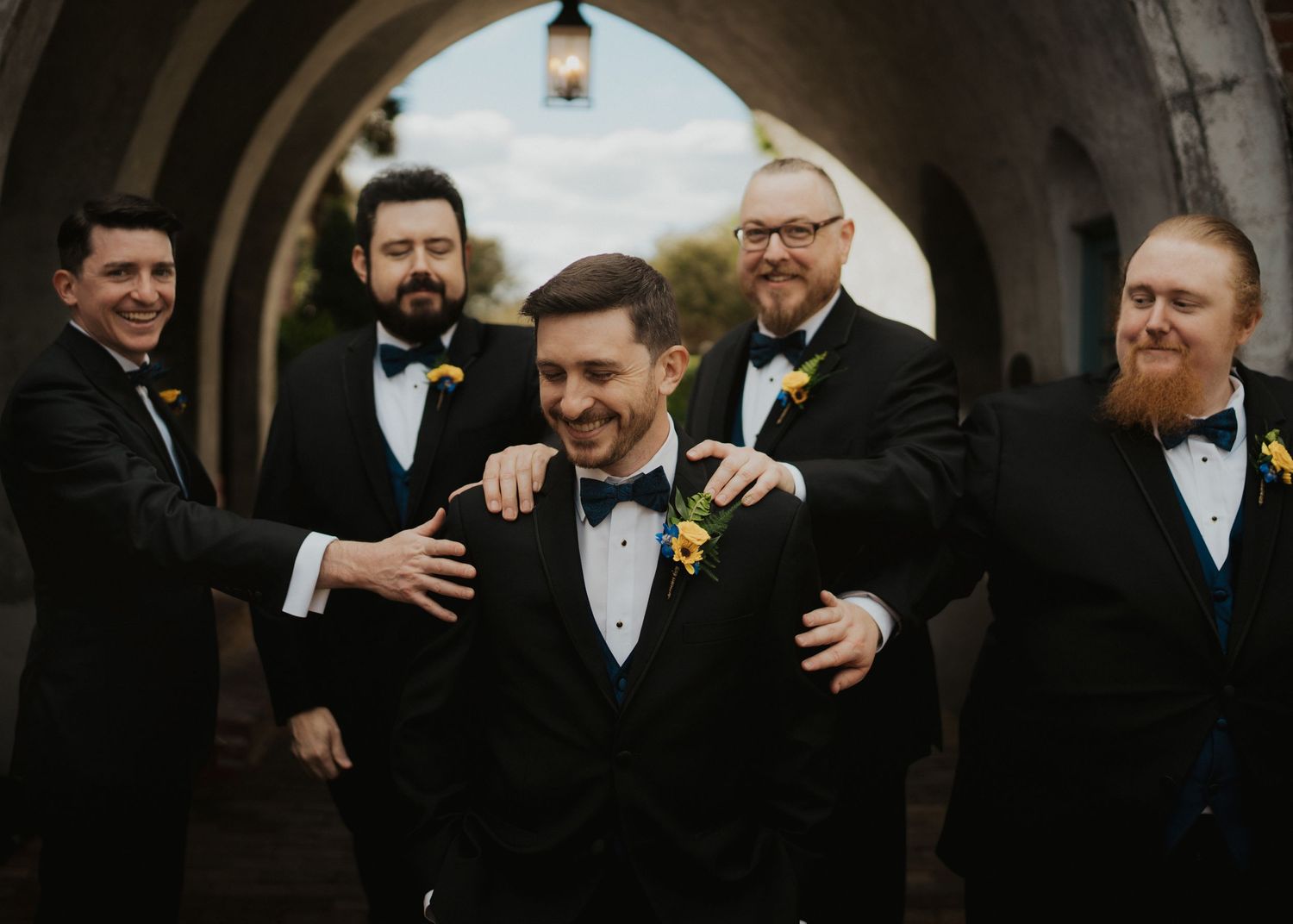 Groomsmen in black tuxedos and bow ties sharing a candid moment in the casa feliz archway