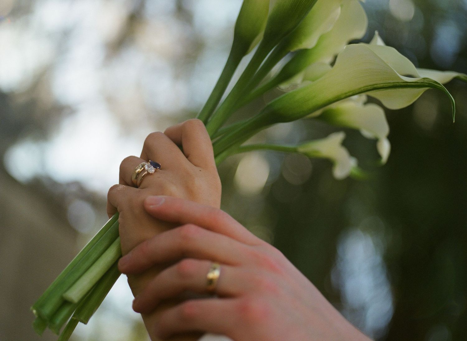 Close-up of hands holding white calla lily bouquet with wedding rings visible.