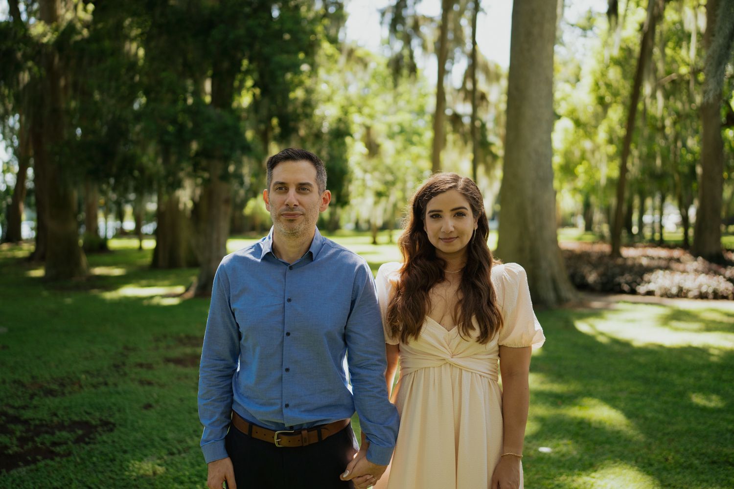A couple poses together in casual formal wear against a backdrop of moss-draped trees.