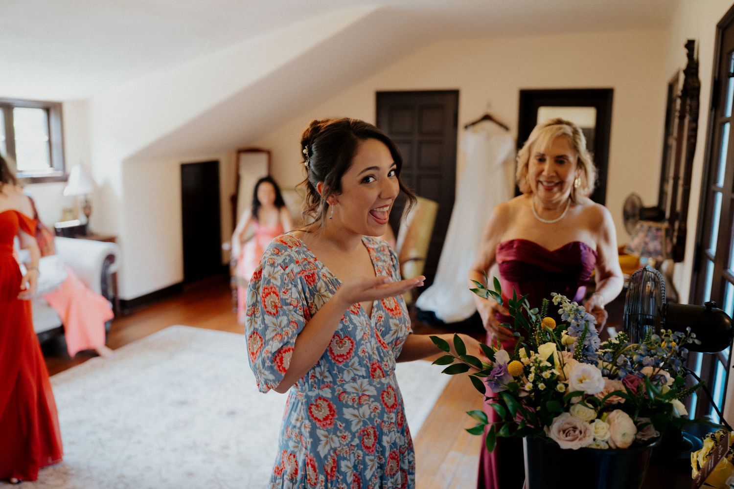 A wedding party gets ready in a bright room with floral dresses and bouquets visible.