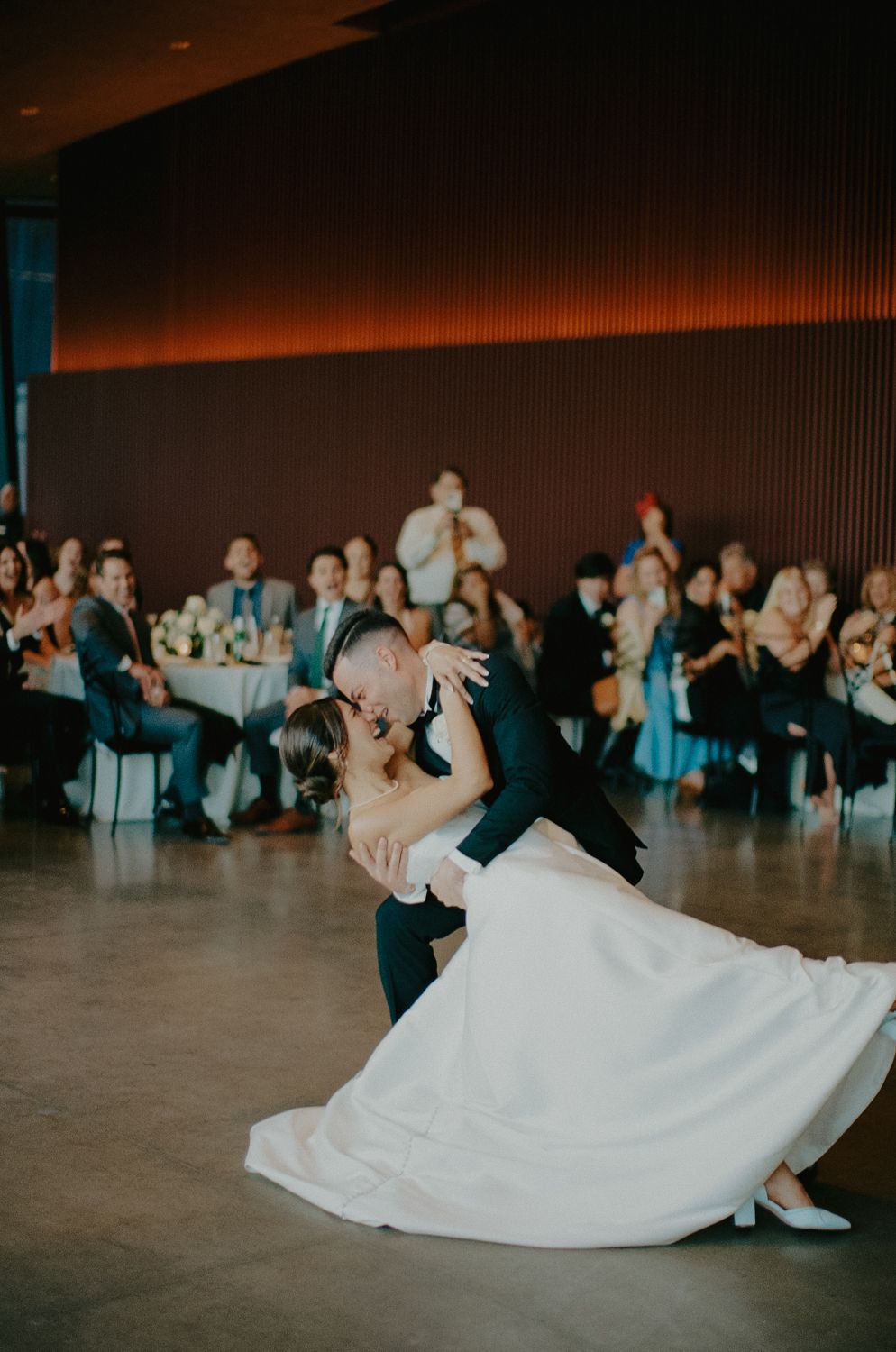 Bride and groom share a romantic dip kiss during their first dance at a wedding reception with guests watching.