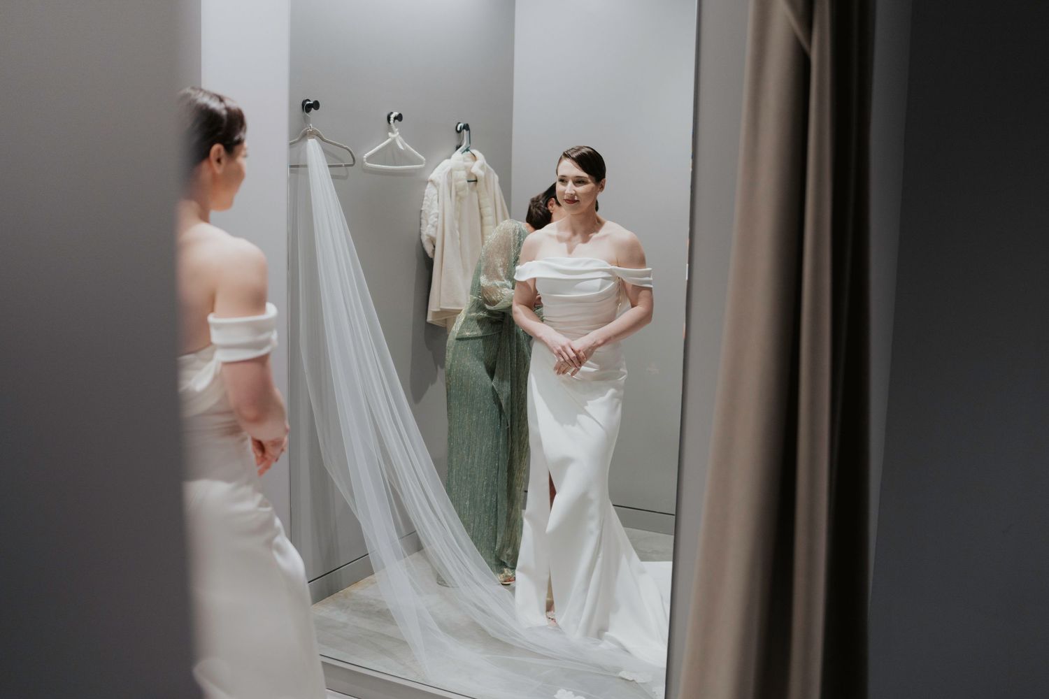 A bride in a classic white wedding gown adjusts her veil while looking in a mirror during pre-ceremony preparations.